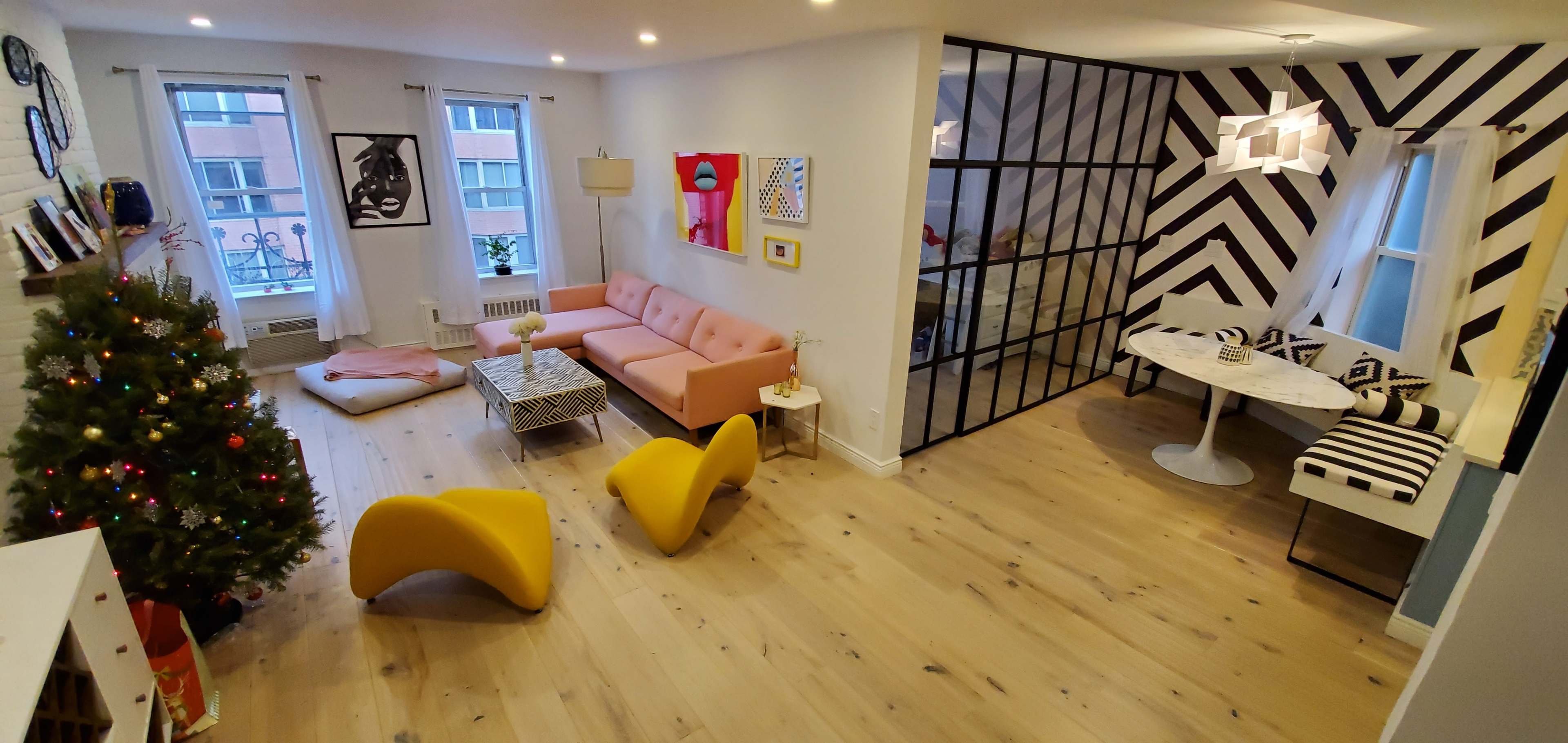 A modern living room with a pink sofa, yellow chairs, a small Christmas tree, and a dining area featuring black and white striped walls.