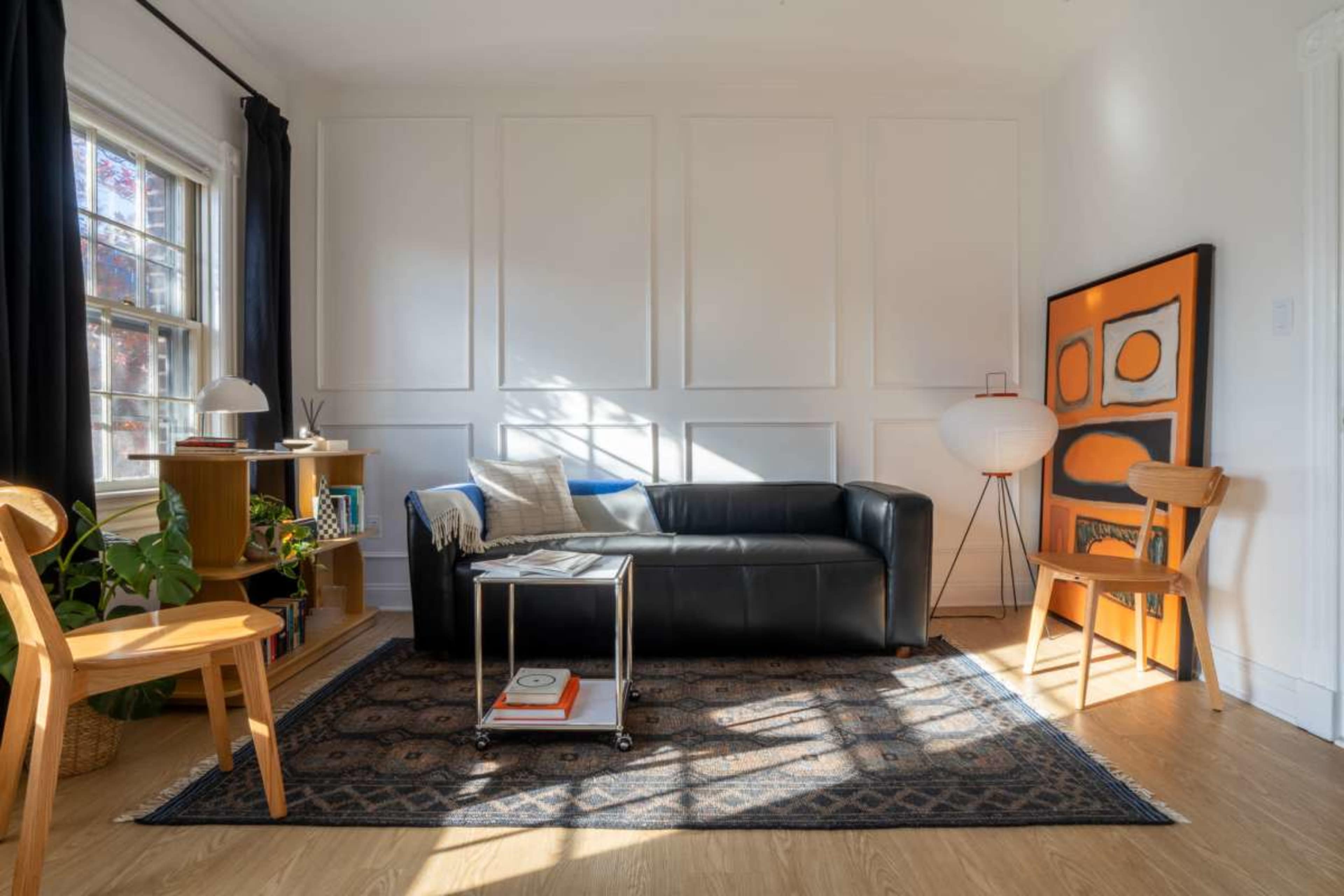 A modern living room features a black leather sofa, a wooden coffee table on wheels, two wooden chairs, a small bookshelf, and a large abstract painting, all illuminated by natural light from a window.
