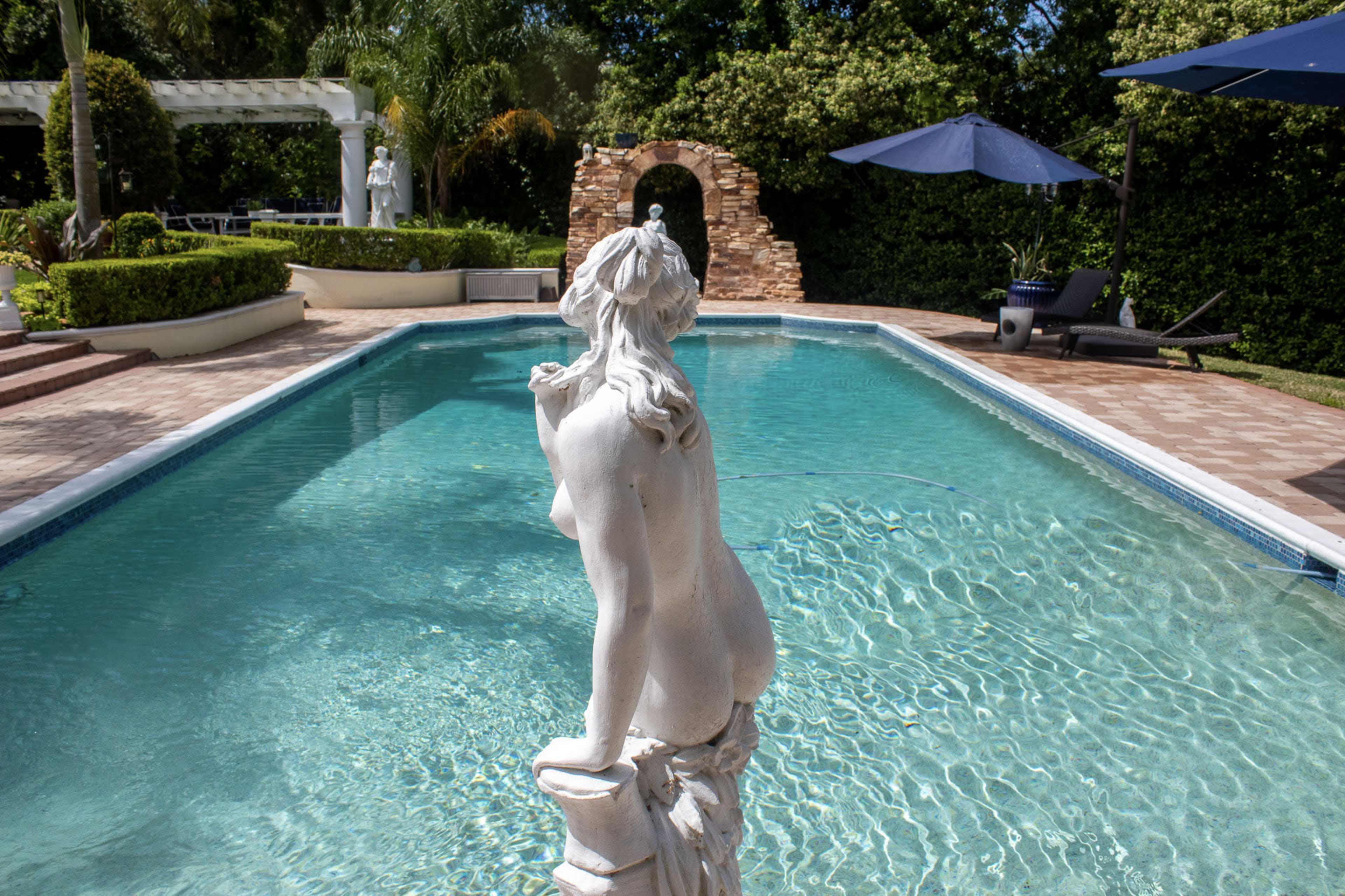 A white statue overlooks a clear swimming pool surrounded by lush greenery and stone features.