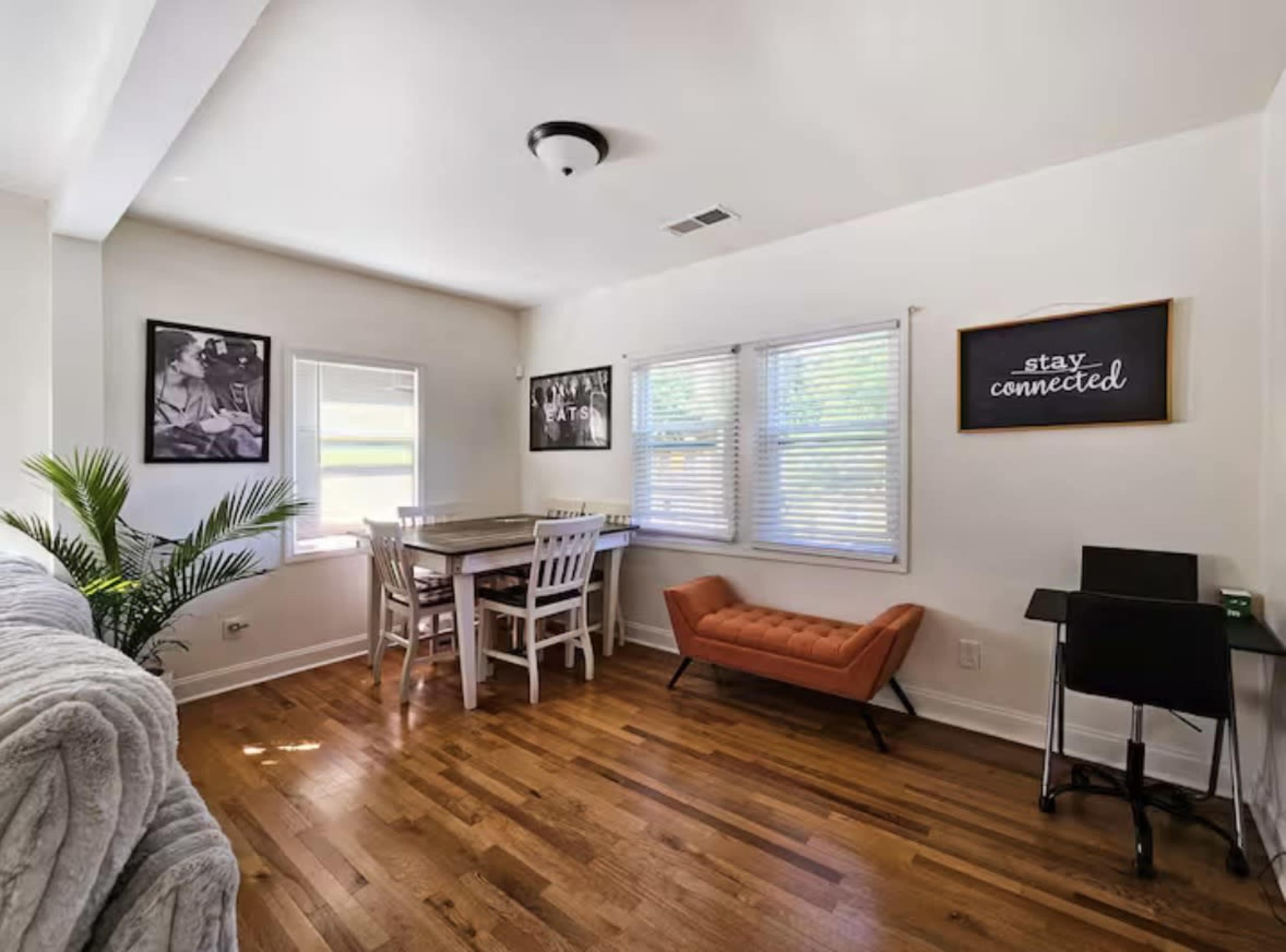 The image features a bright, modern living space with a dining table and chairs, a sofa, a decorative orange bench, and framed artworks on the walls.