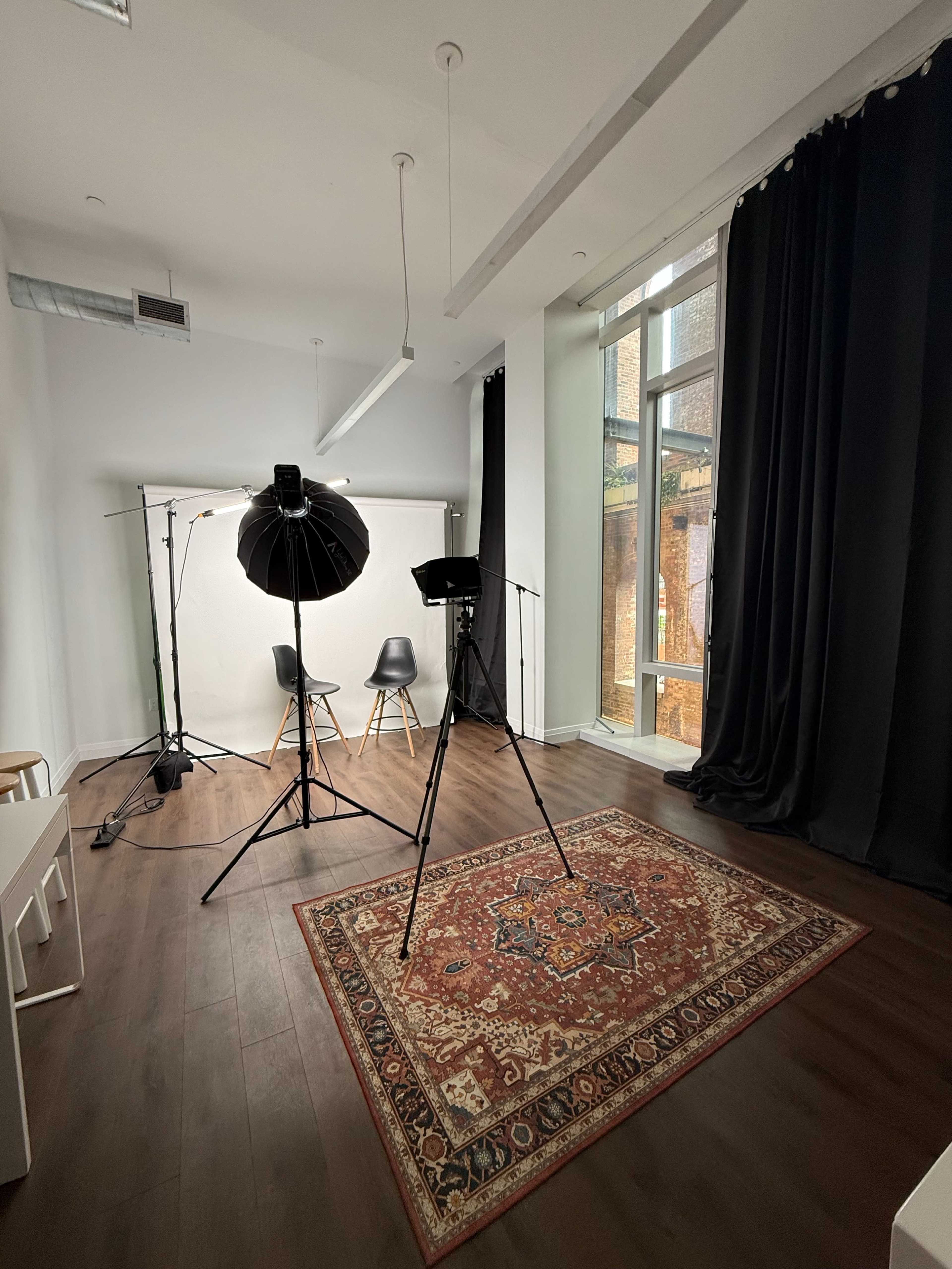 The image shows a photography studio with a light setup, two chairs, a backdrop, and a large window allowing natural light to enter.