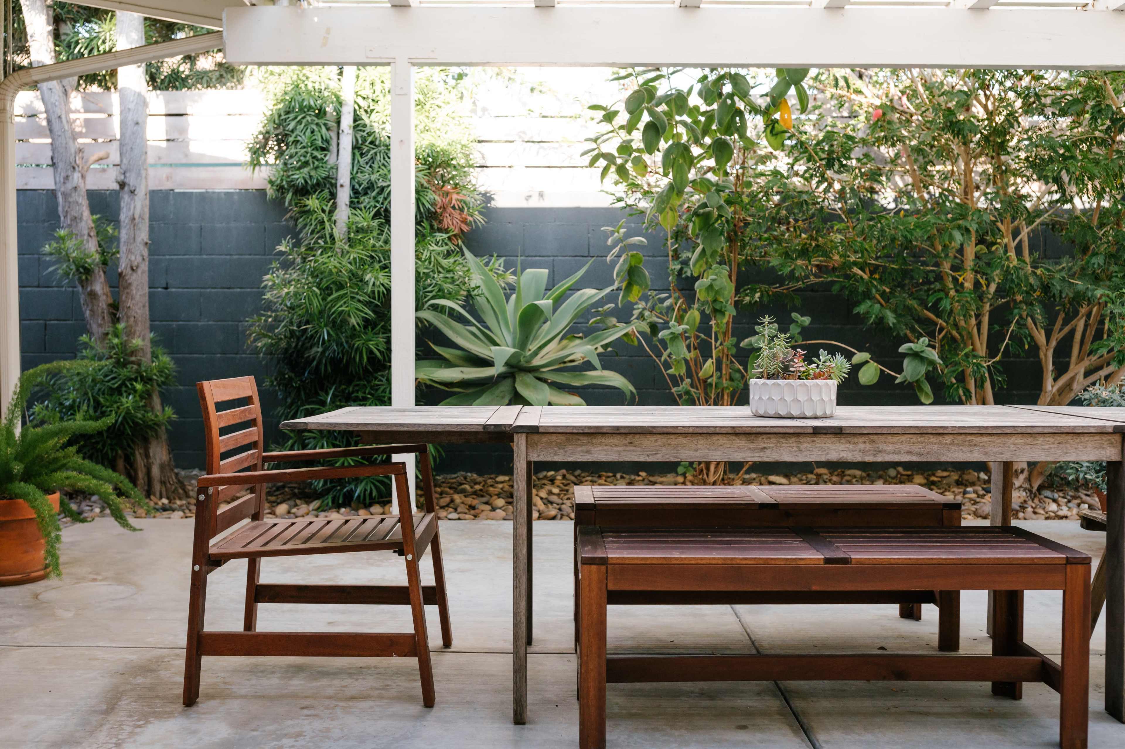 A wooden table and chairs are set under a covered patio, surrounded by greenery and a stone wall.