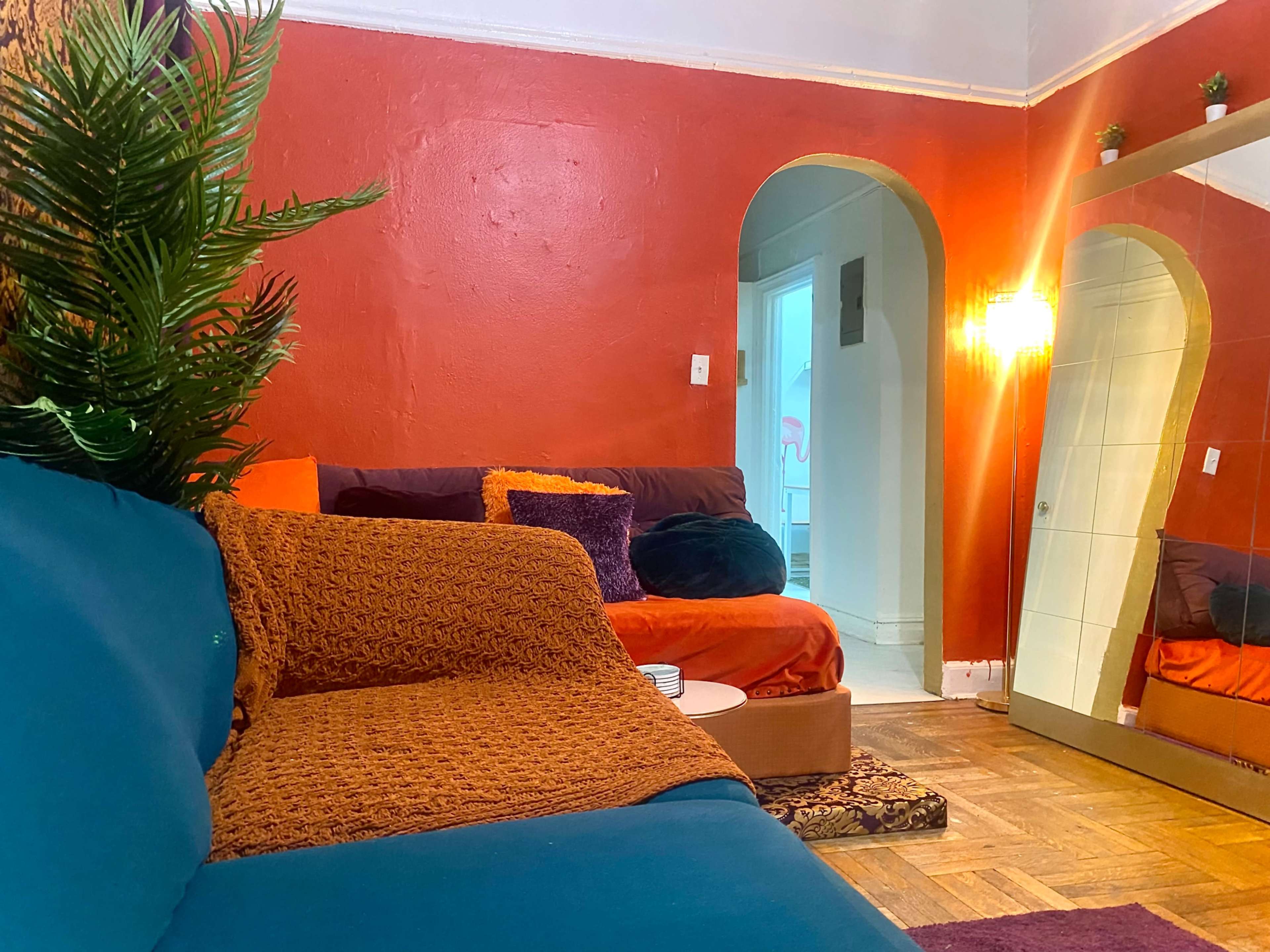 The room features a blue couch, an orange wall, and a large mirror reflecting a cozy seating area with various cushions and a potted plant.