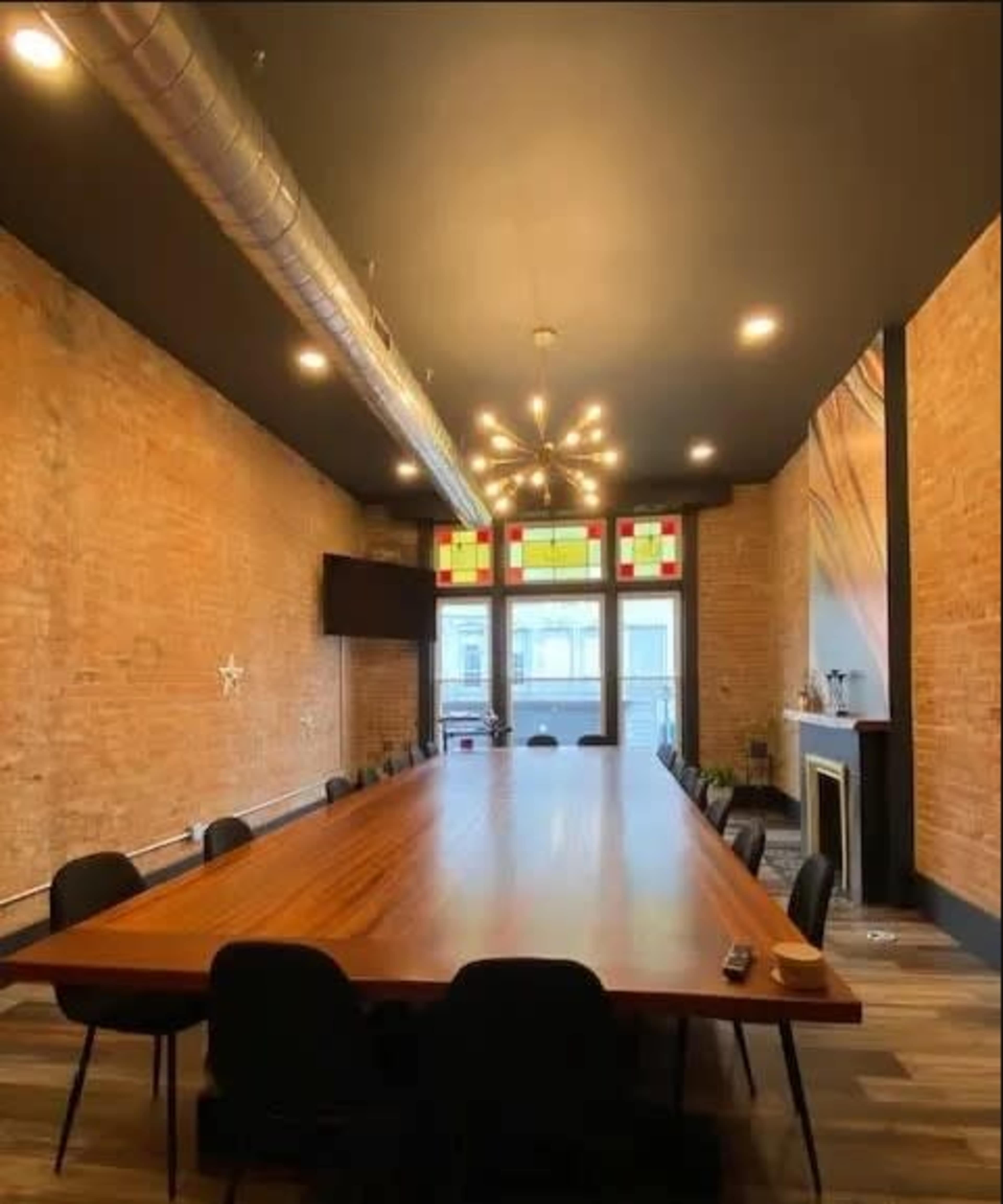 A spacious conference room features a long wooden table, exposed brick walls, and large windows with colorful panels.