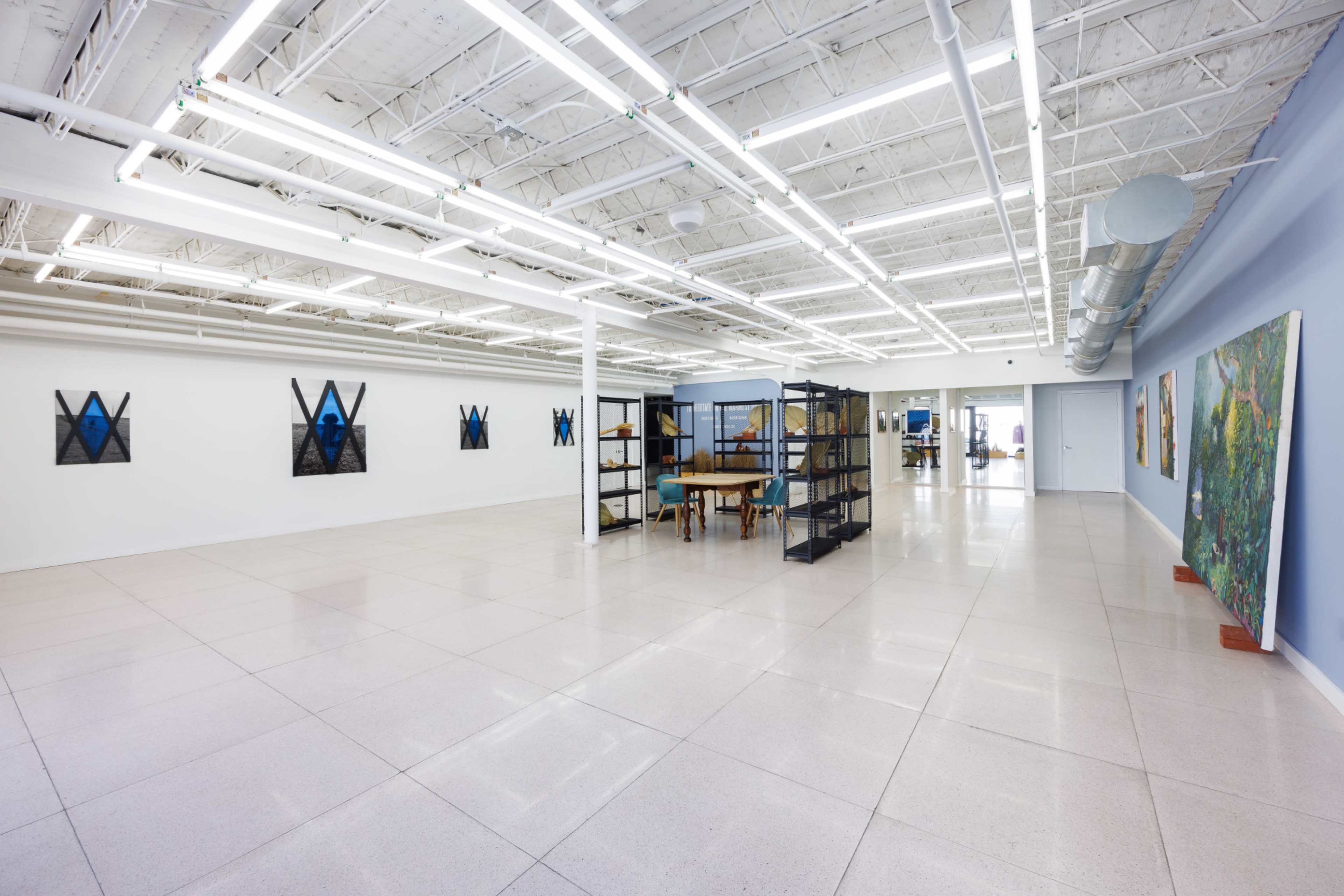 The image shows a spacious, well-lit interior of an art gallery featuring minimalist decor, large windows, and artwork displayed on the walls.