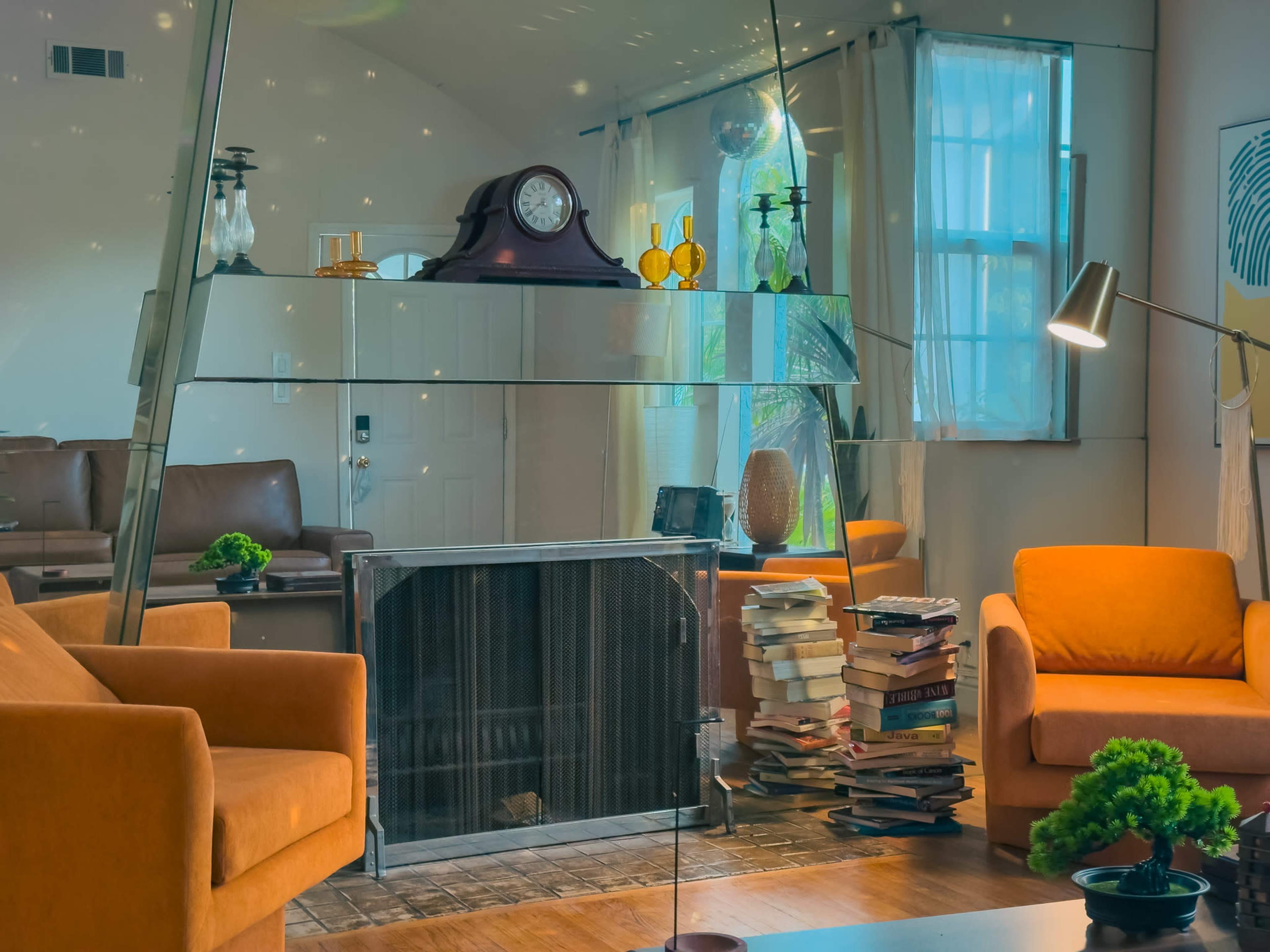 The image shows a modern living room with orange armchairs, a glass shelf displaying decorative items, a fireplace, and a stack of books beside it.