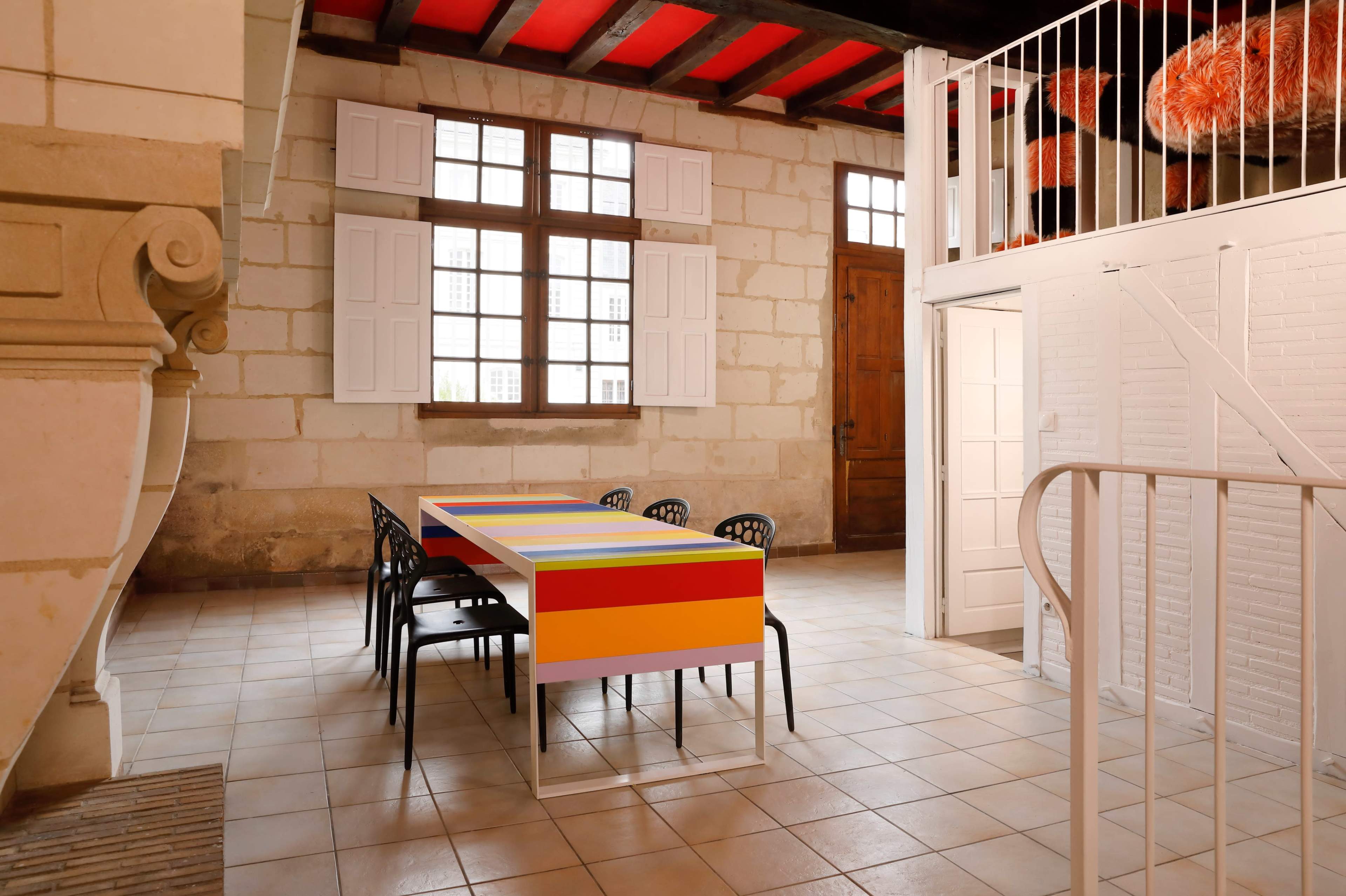 The image shows a spacious room with a long, colorful table surrounded by black chairs, wooden beams on the ceiling, and large windows with white shutters.