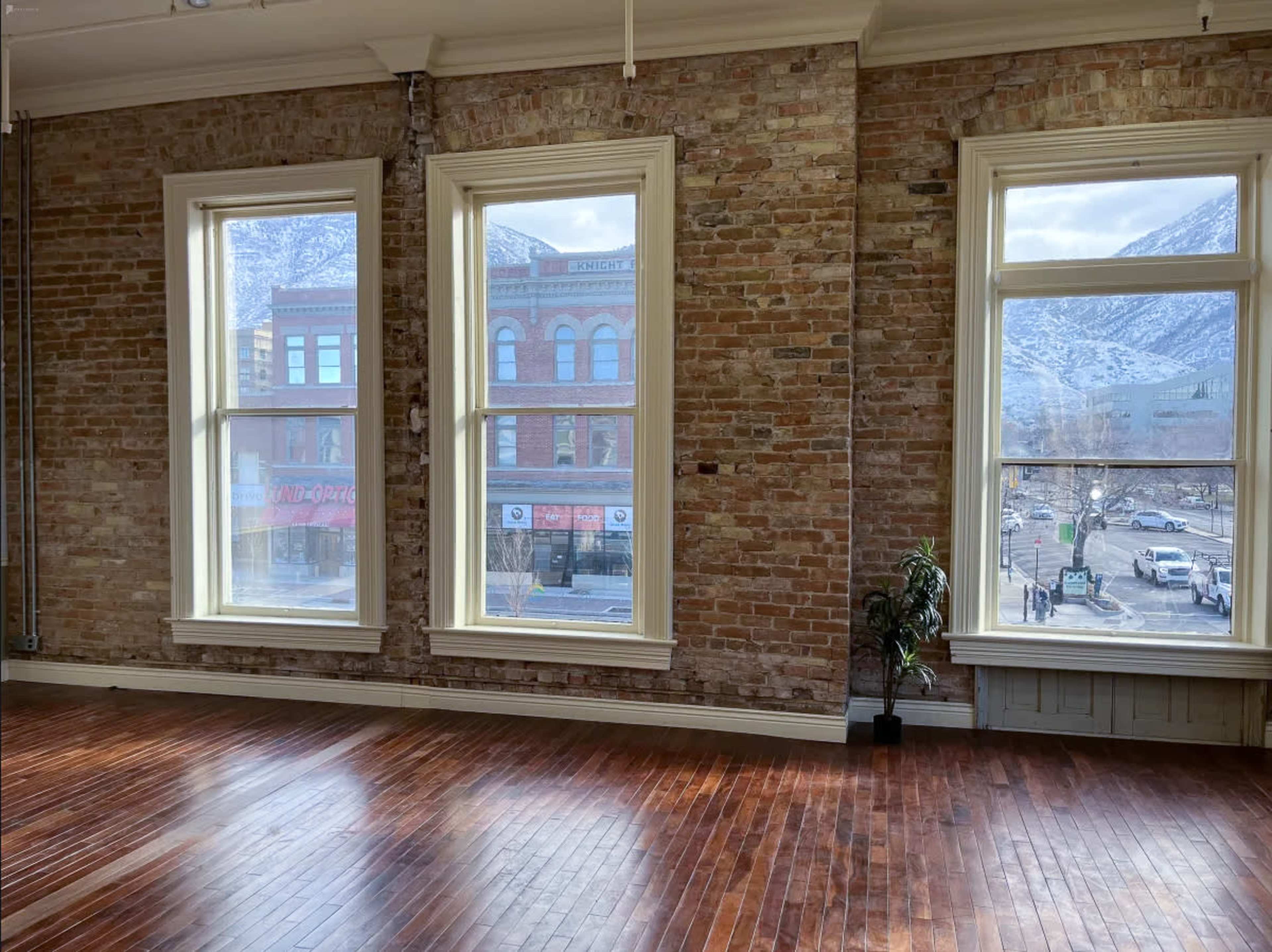 The room features large windows with a view of a street and mountains, alongside exposed brick walls and wooden flooring.