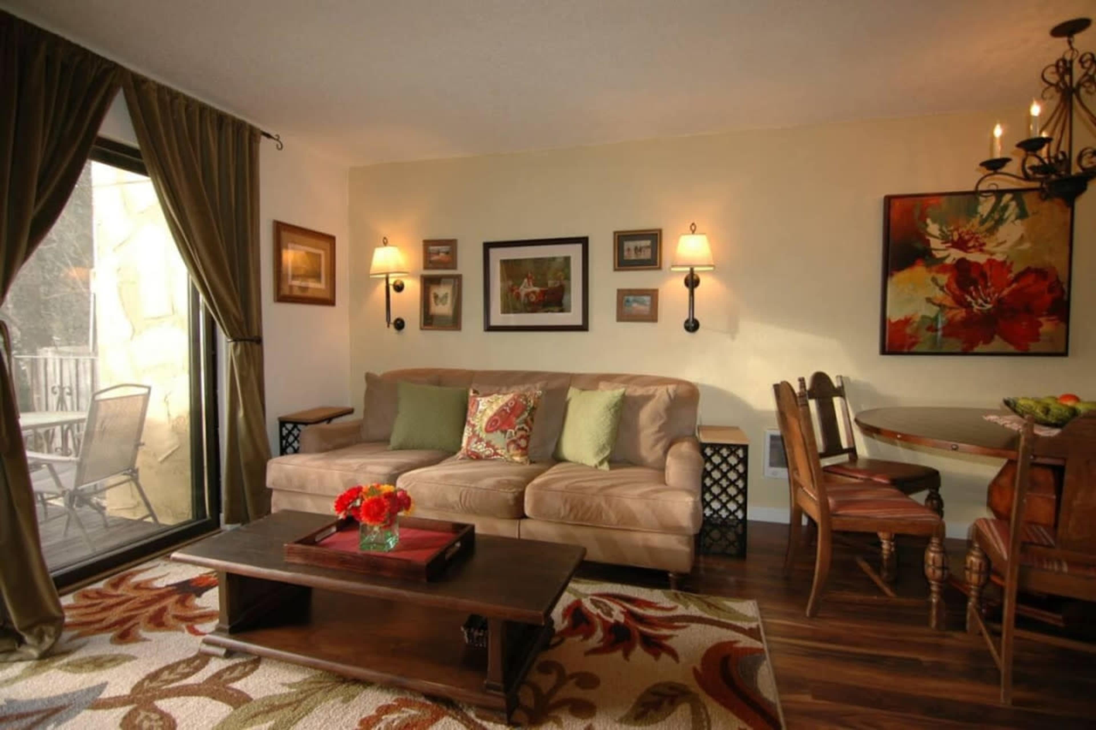 The image shows a cozy living room with a beige sofa, decorative pillows, a wooden coffee table, and a dining area with a table and chairs.