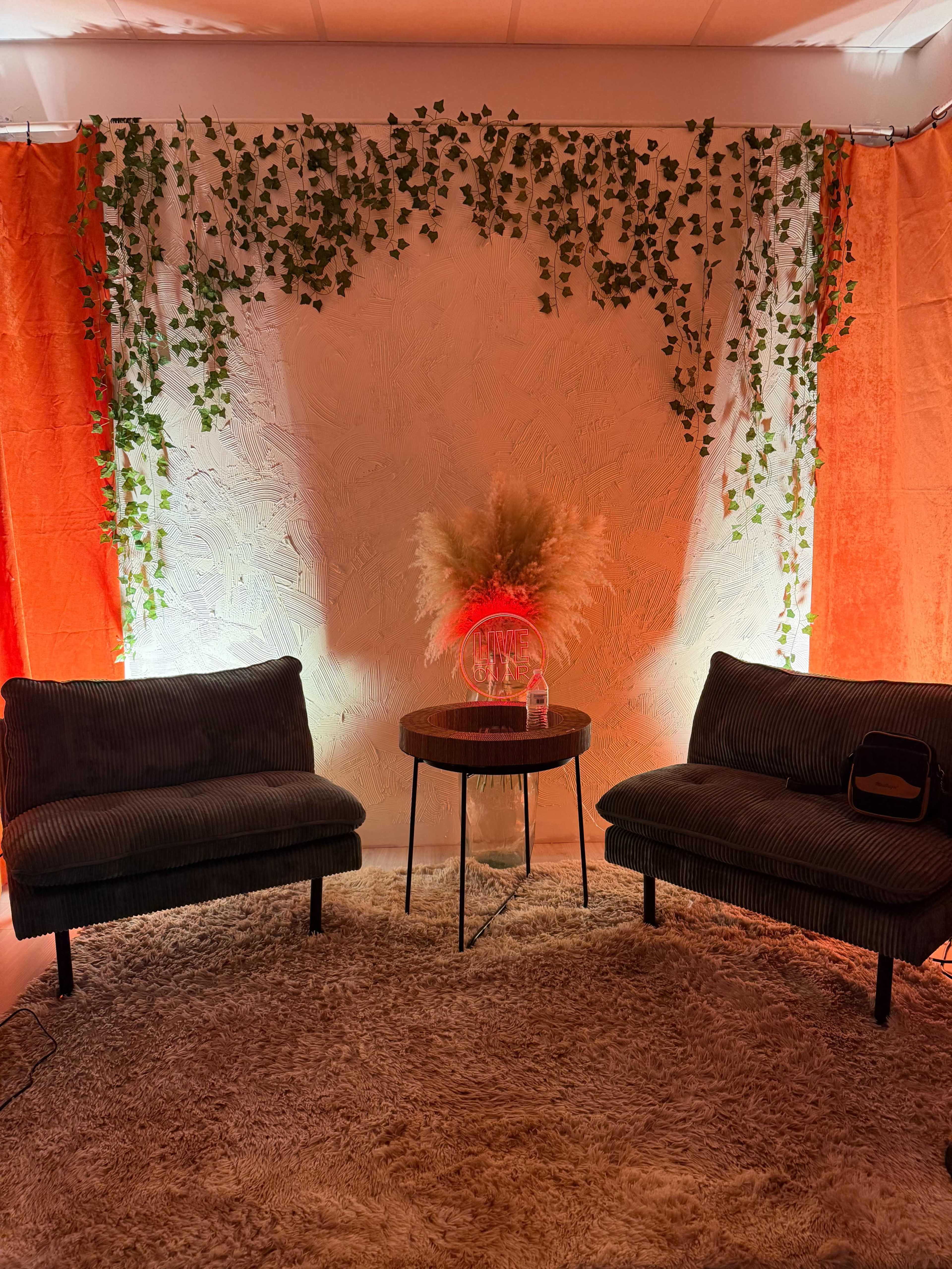The scene features two upholstered chairs facing a small circular table surrounded by a decorative backdrop of ivy and vibrant orange curtains.