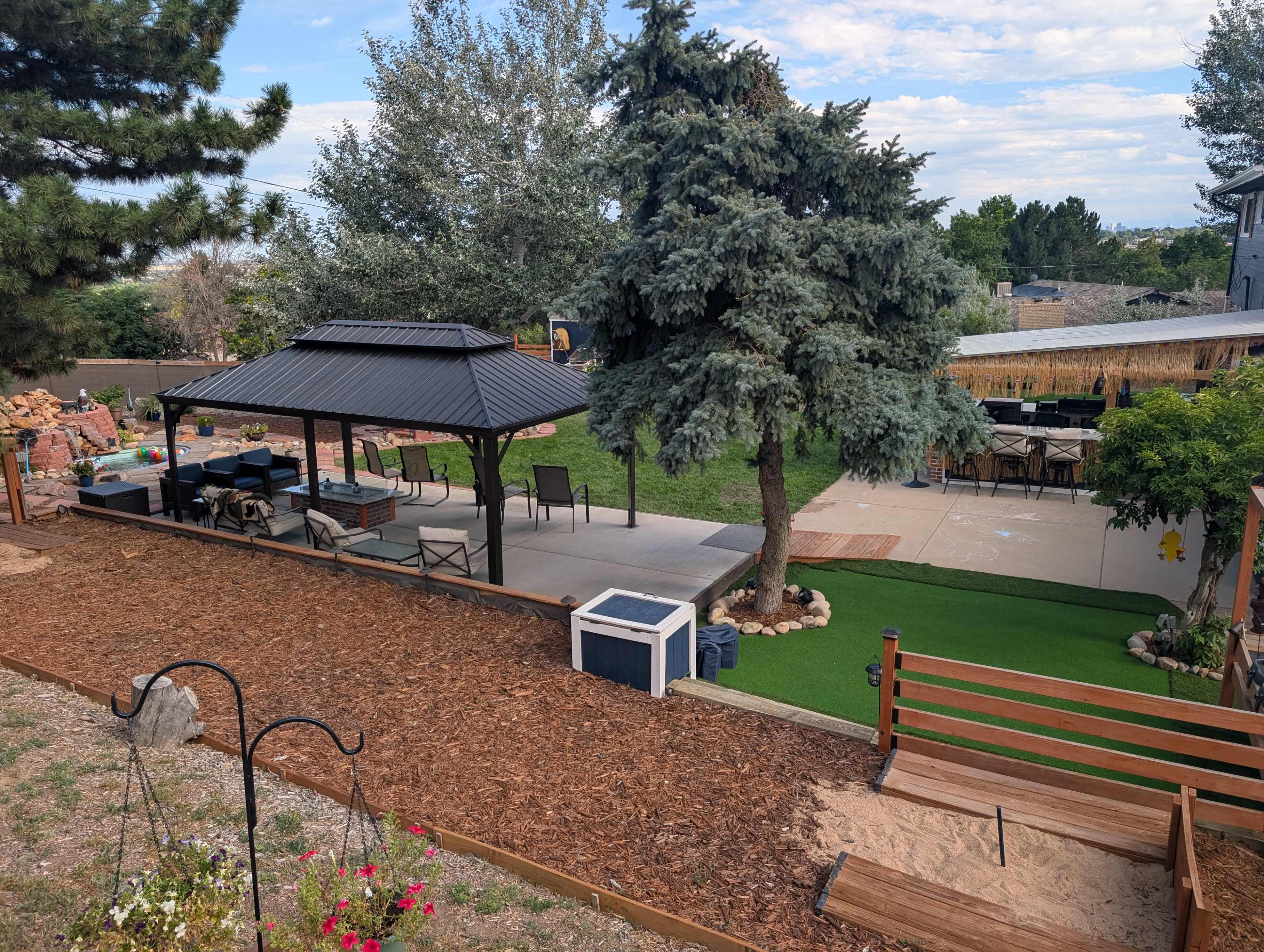 The image shows a backyard with a gazebo, outdoor seating area, and a green lawn, surrounded by trees and landscaped features.