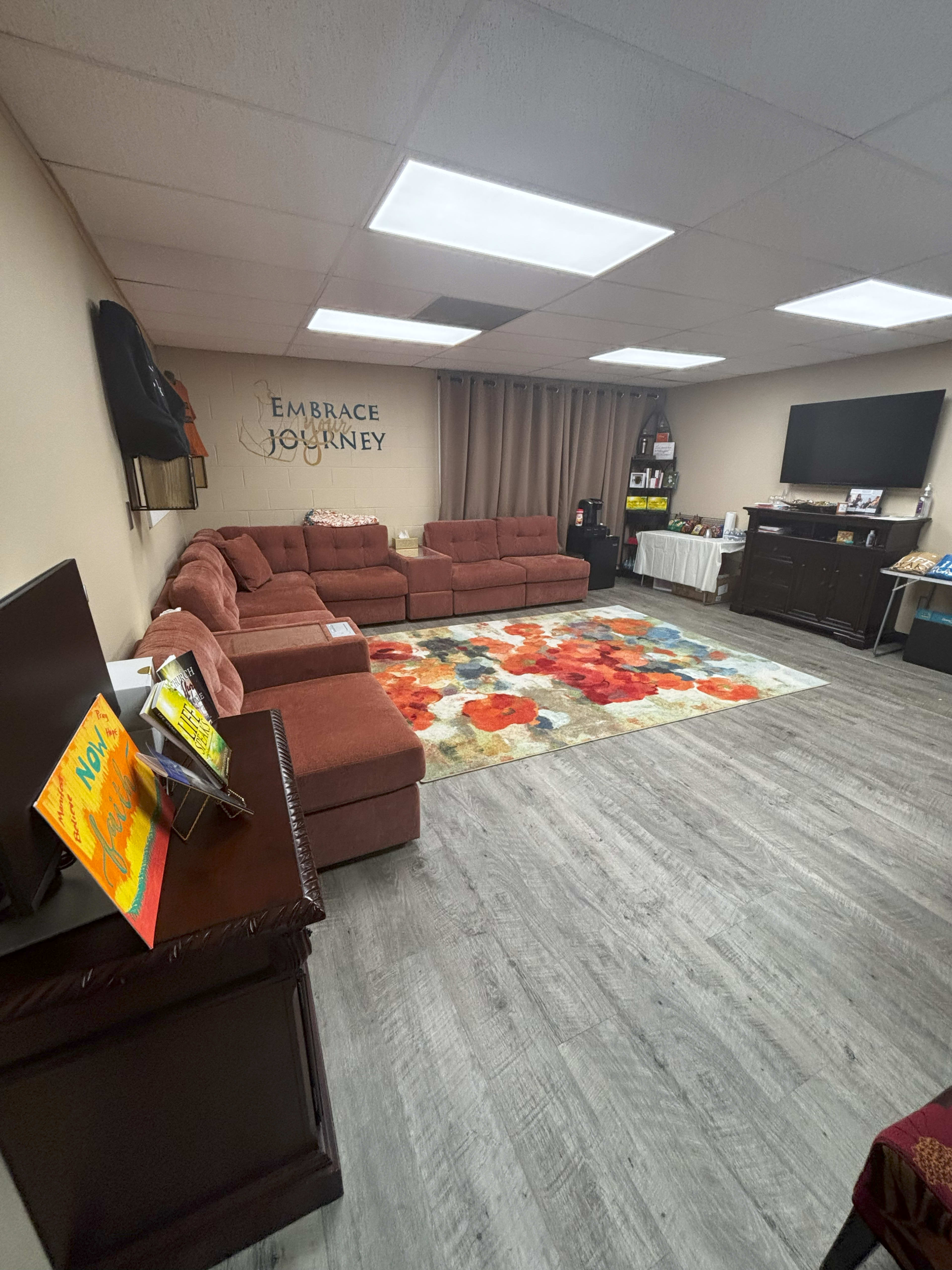The image shows a cozy multi-purpose room featuring a large, L-shaped sectional sofa, a colorful area rug, a television, and shelves with various items.