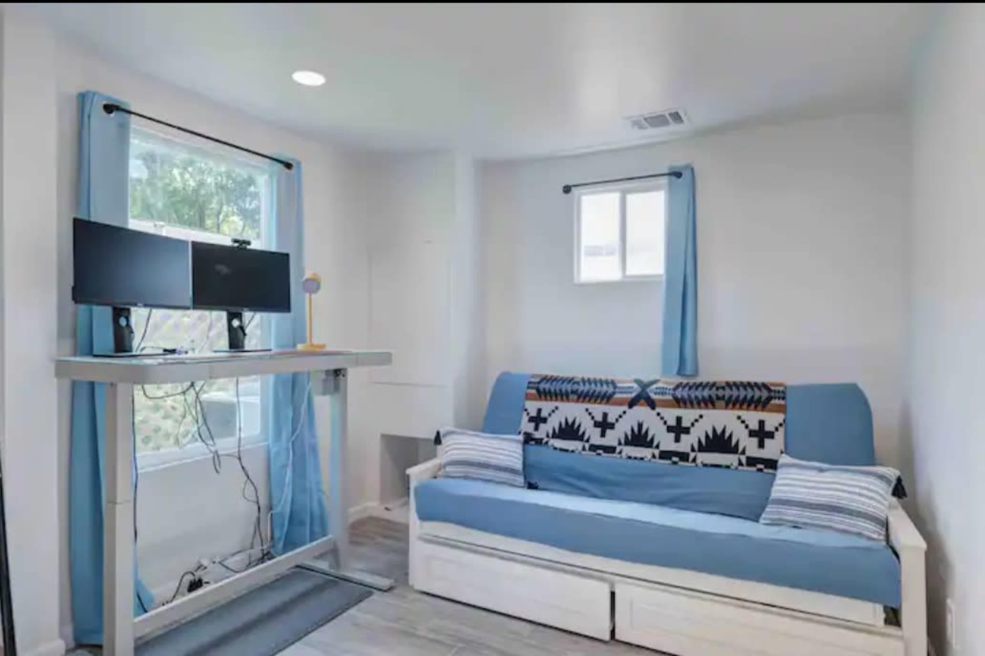 The room features a blue sofa with a patterned cover, a standing desk with dual monitors, and light blue curtains covering the windows.