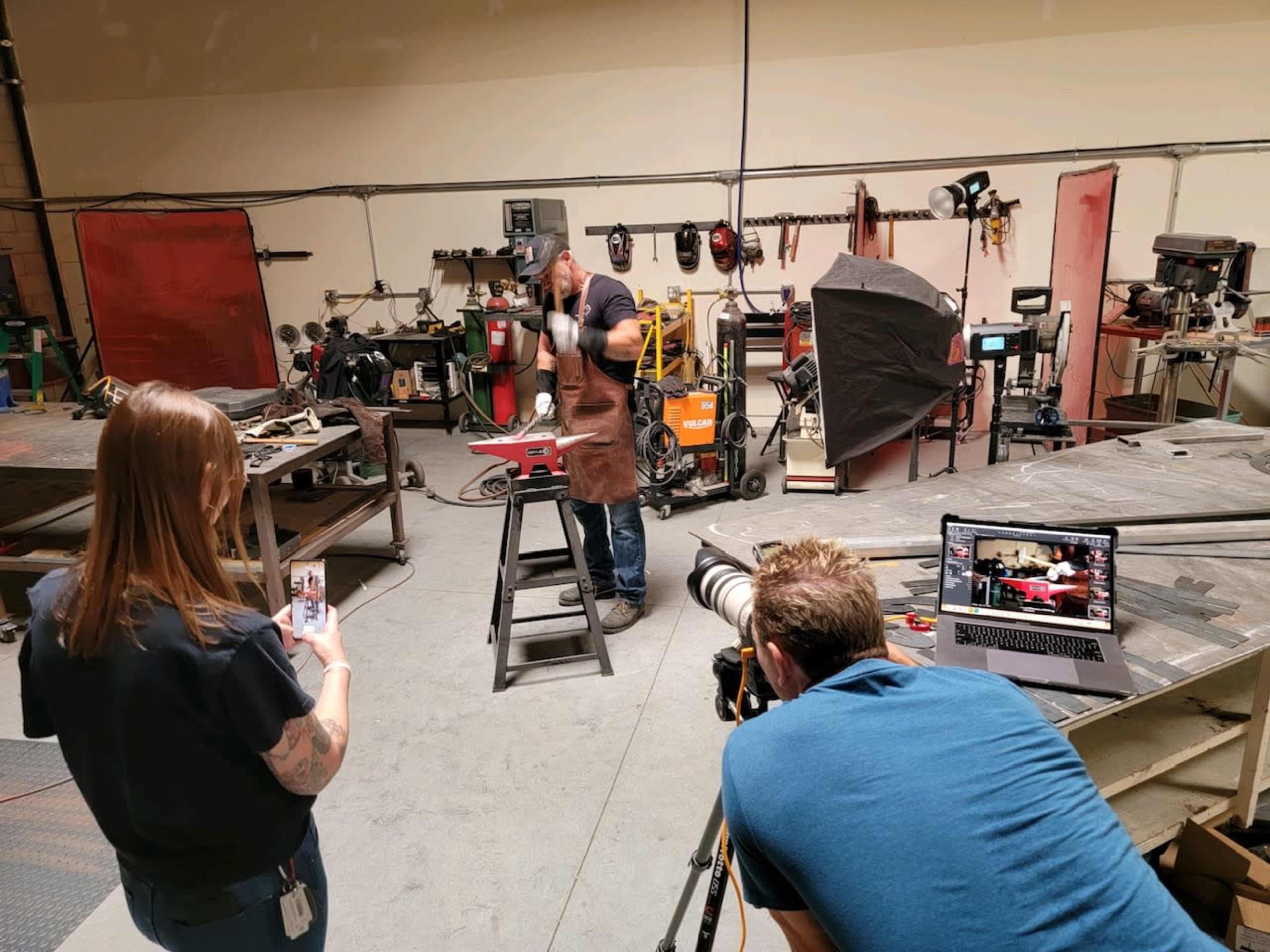 A person wearing an apron works on a metal project in a workshop while another person records on a phone and a third person operates a camera.