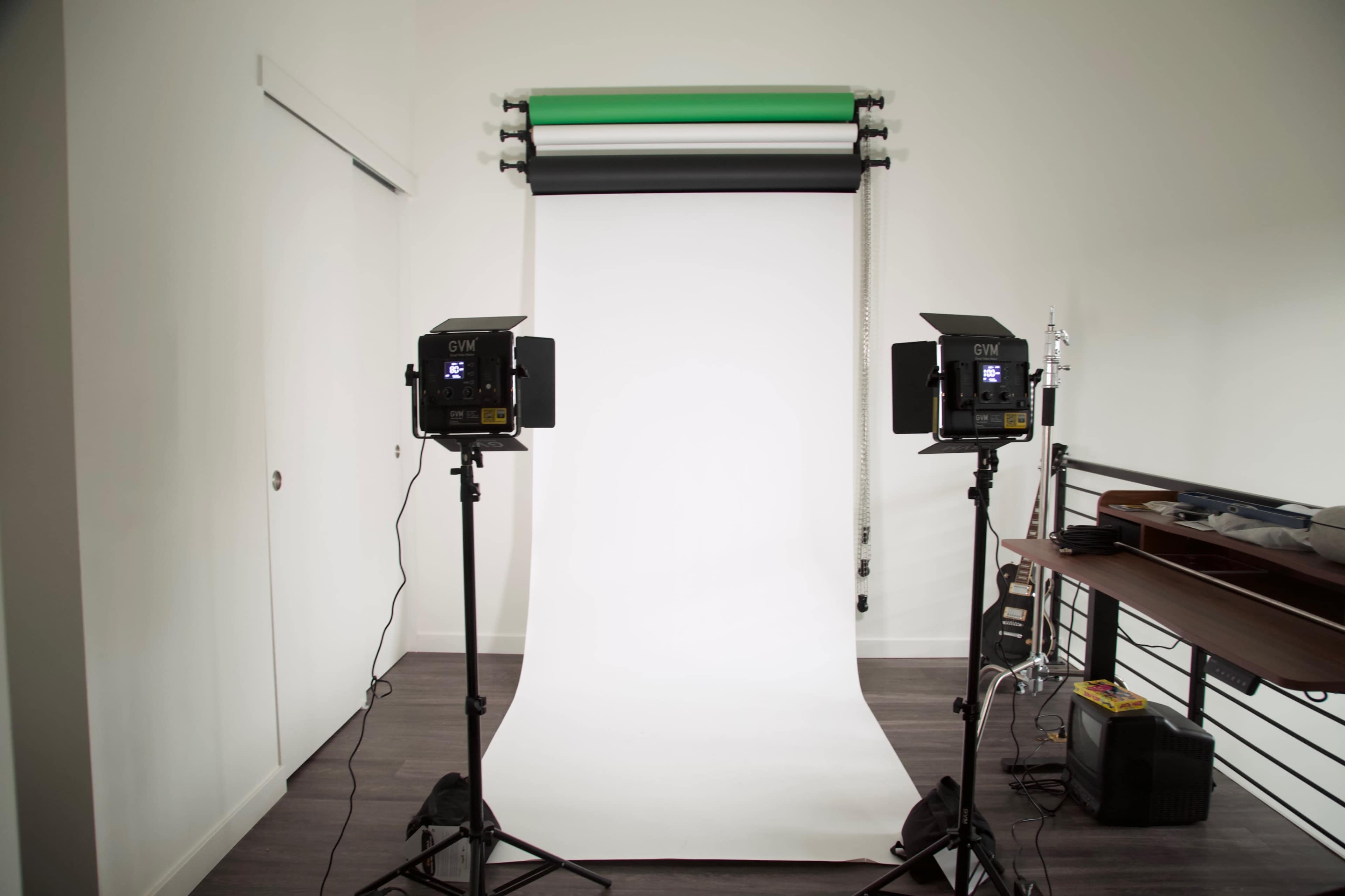 A well-lit photography studio setup features two lights on stands facing a blank white backdrop with green and black rolls above.