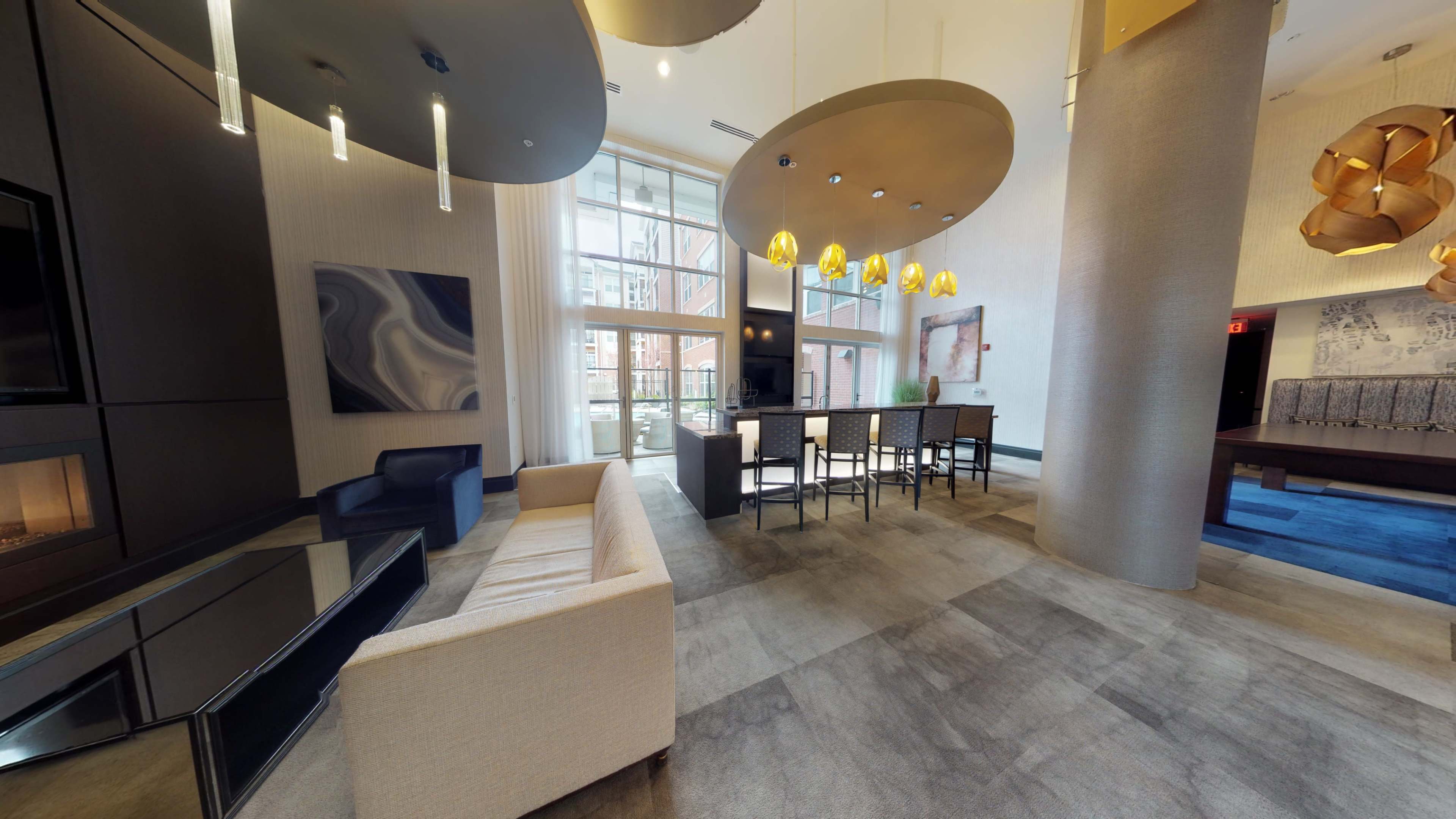 A modern lounge area featuring a light-colored sofa, a bar with high chairs, and large windows allowing natural light to enter.