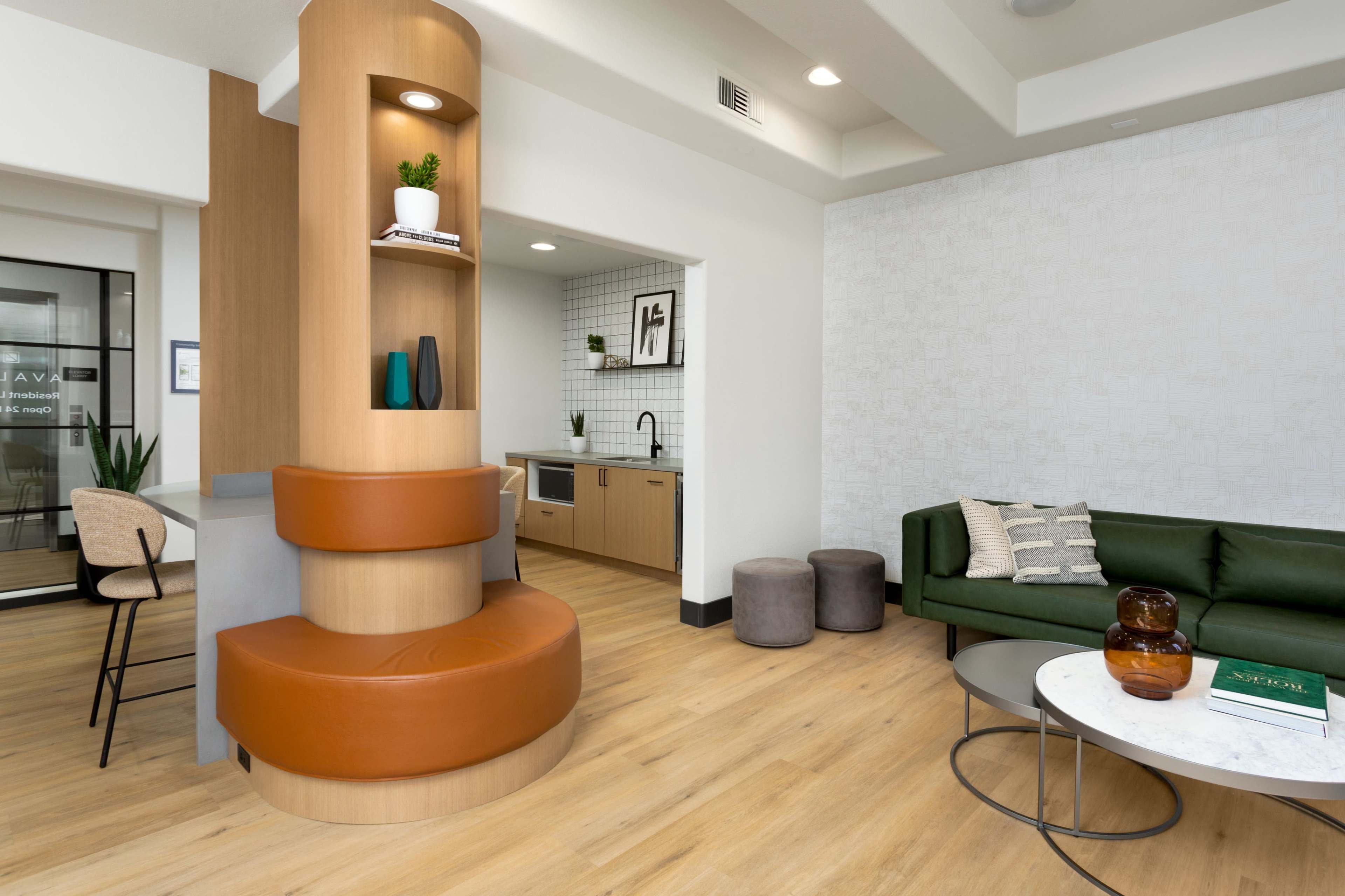 A modern lounge area with a kitchenette, featuring wood accents, green seating, and a circular counter with brown upholstery.