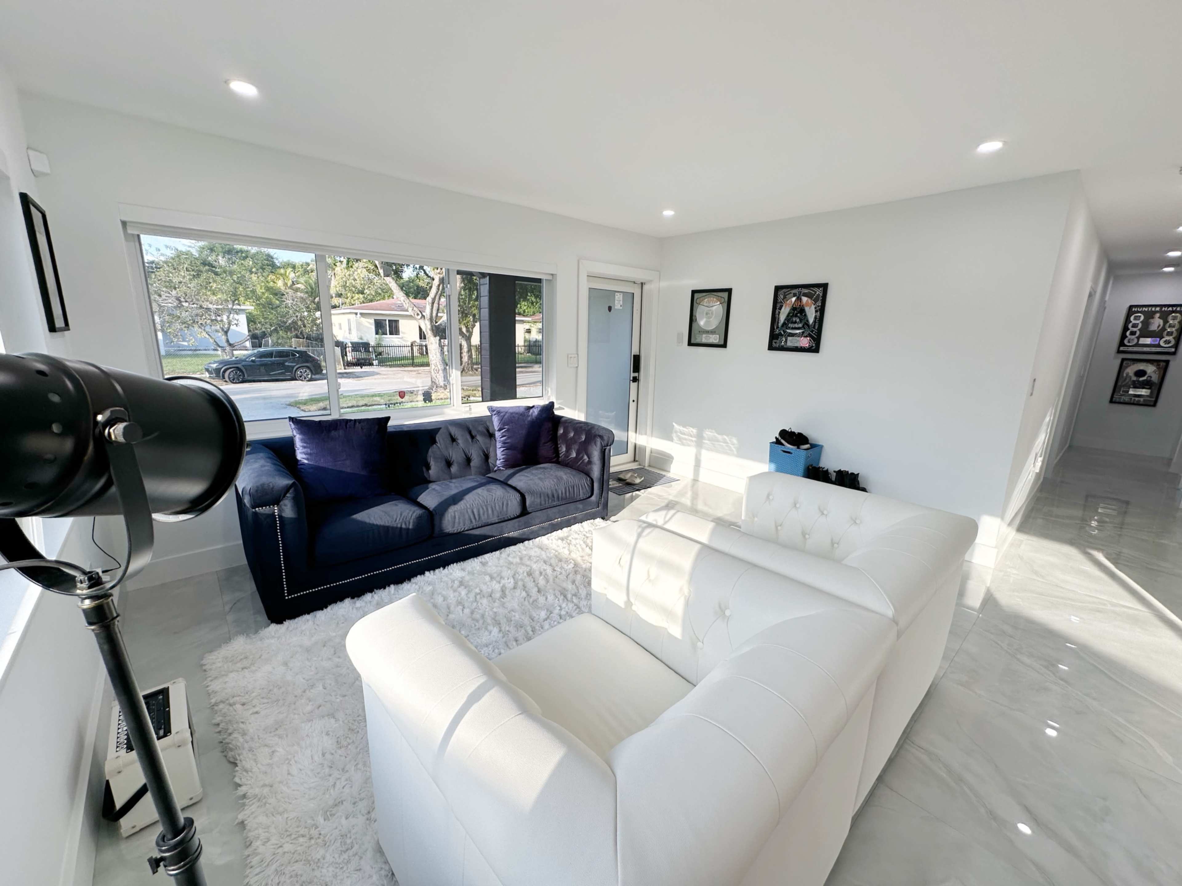 The living room features a white chesterfield sofa, a dark blue couch, and a shag rug, all illuminated by natural light from large windows.