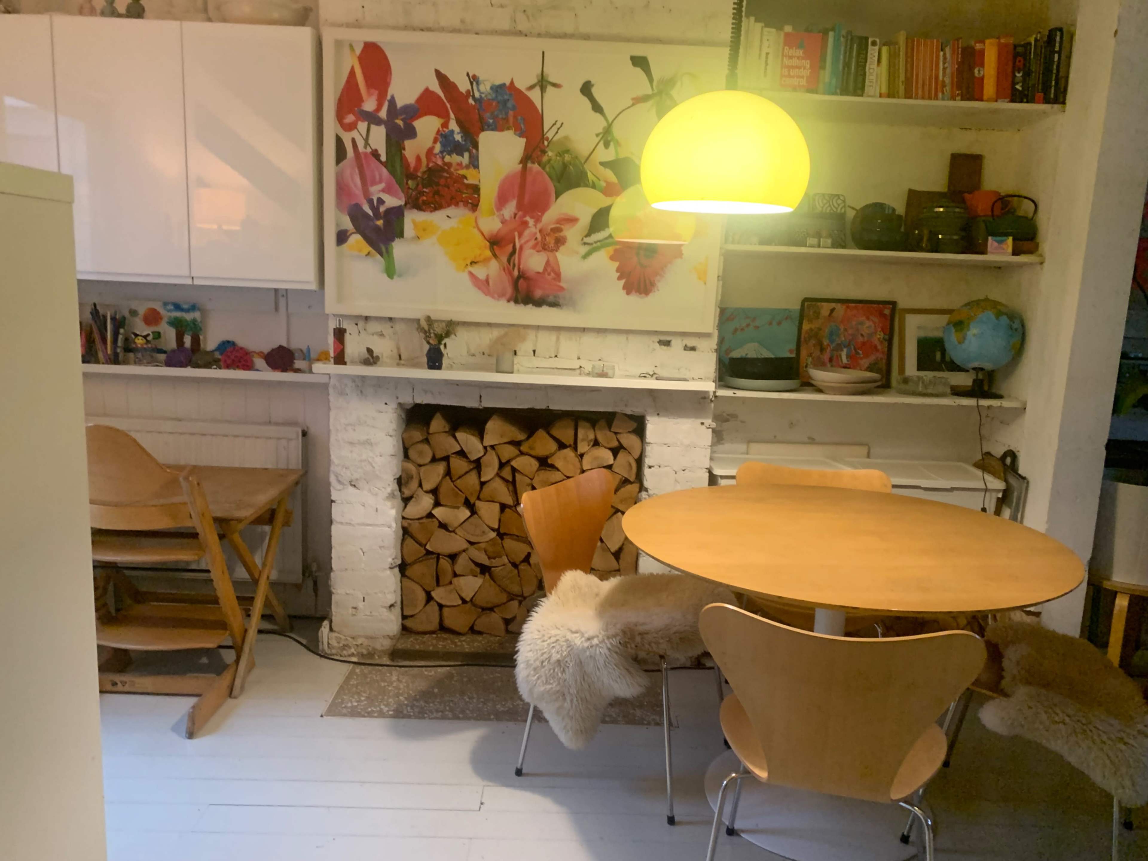 The image shows a cozy kitchen or dining area with a round wooden table, wooden chairs, a white brick fireplace stacked with logs, and colorful artwork on the wall.