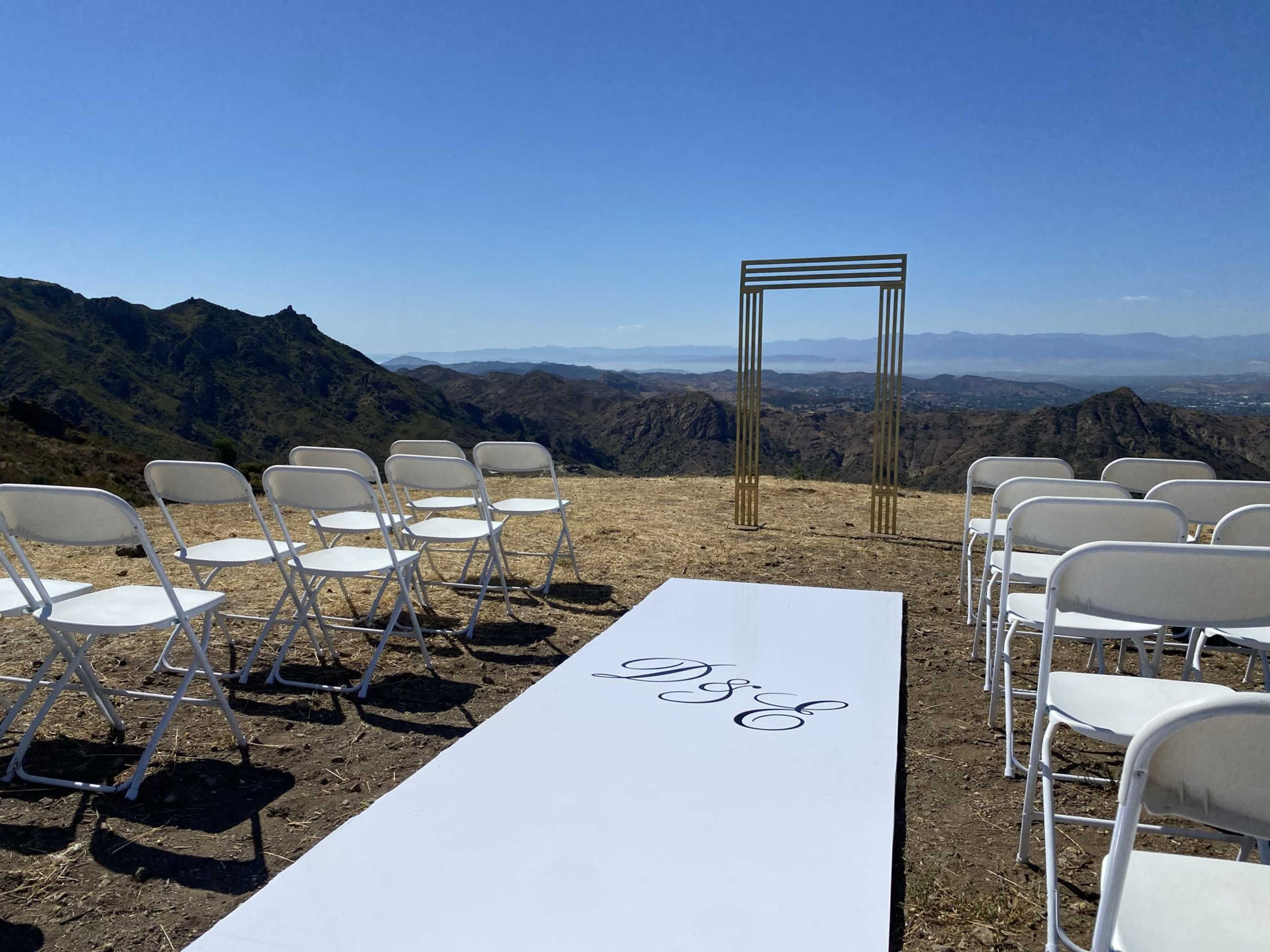 A wedding ceremony setup features white chairs arranged on a grassy area with a mountain backdrop and a decorative arch.