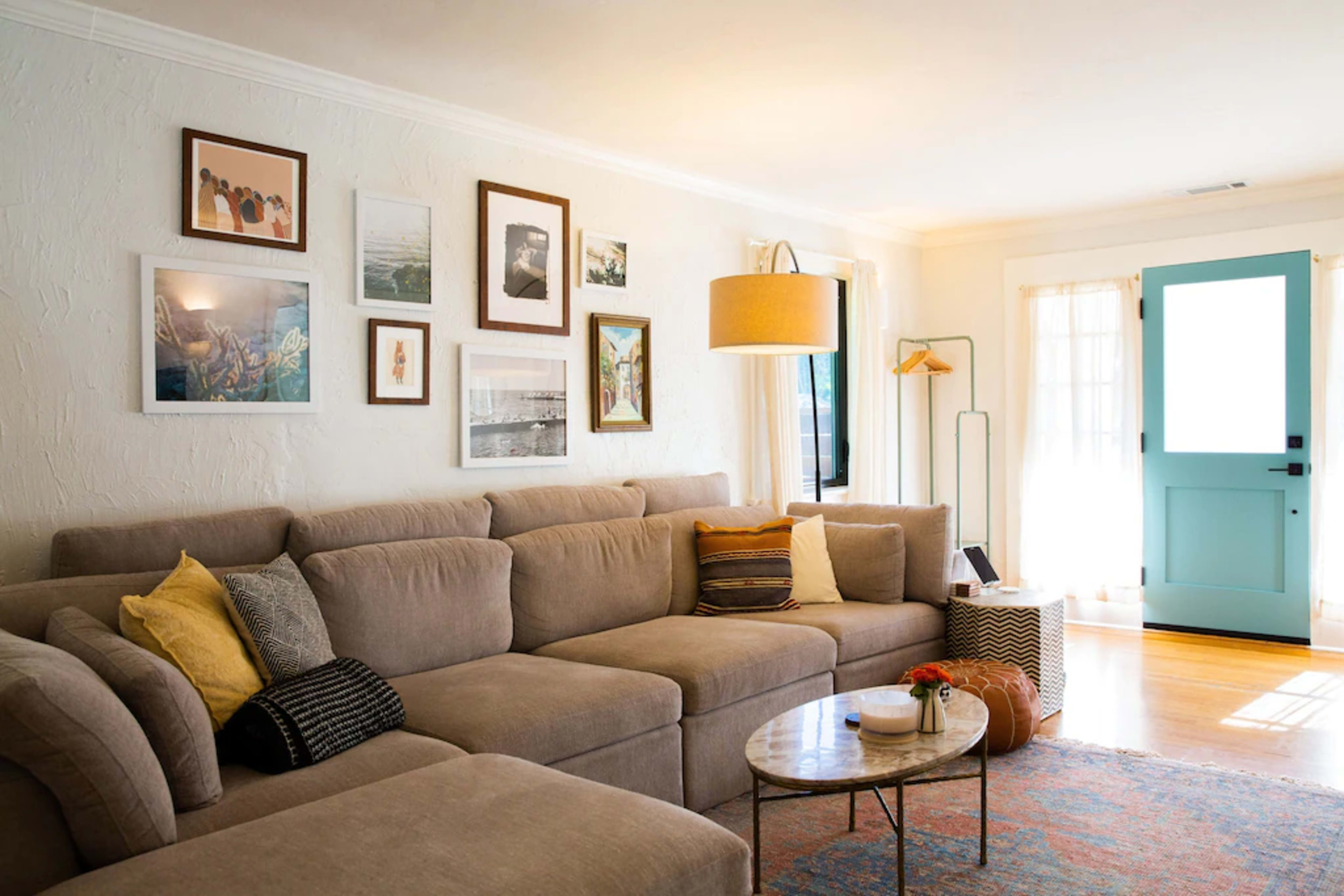 The living room features a large beige sectional sofa arranged around a round coffee table, with framed artwork on the walls and a light blue front door visible in the background.