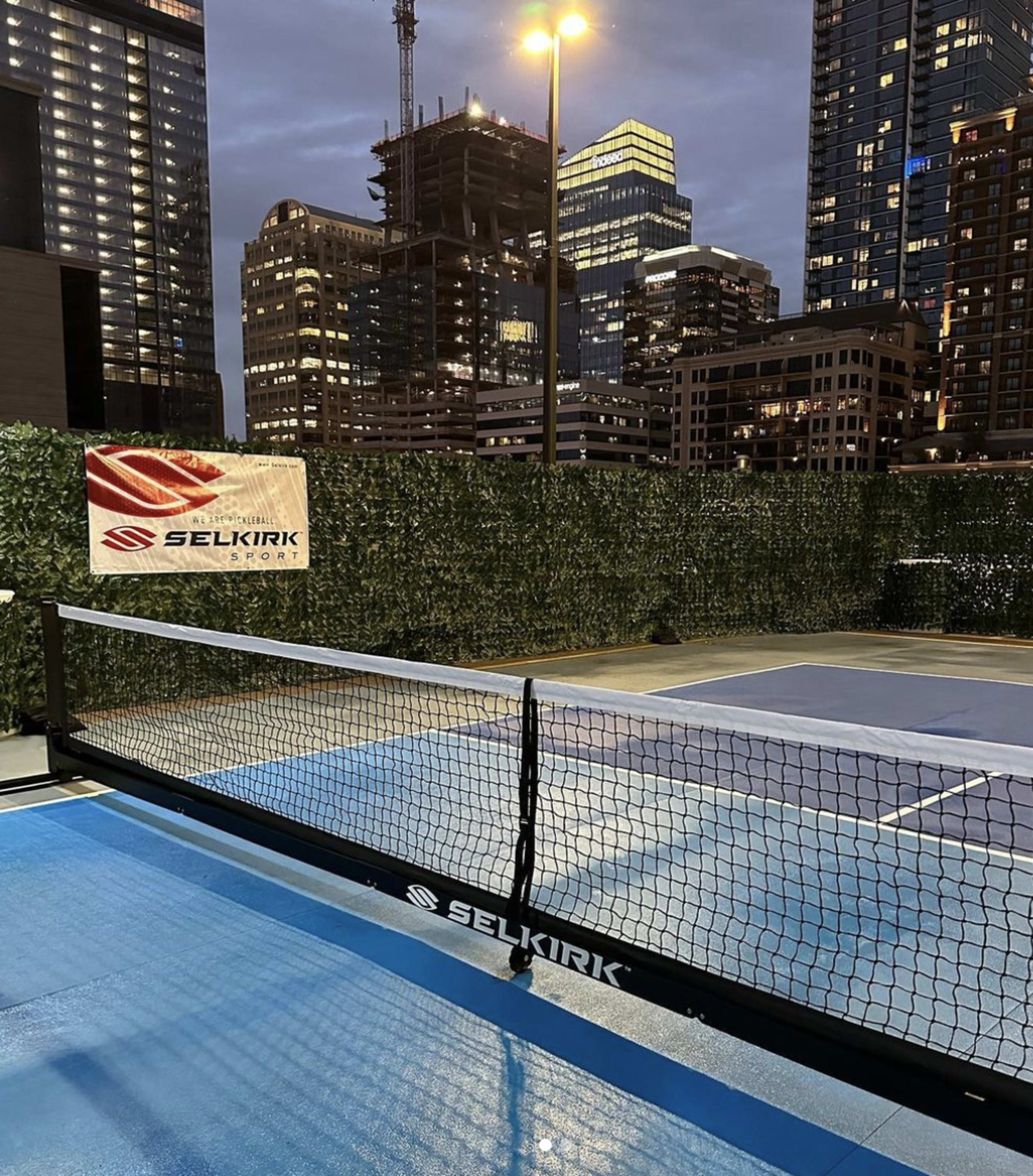 A brightly lit pickleball court with a net and surrounding greenery, set against a backdrop of urban buildings at dusk.