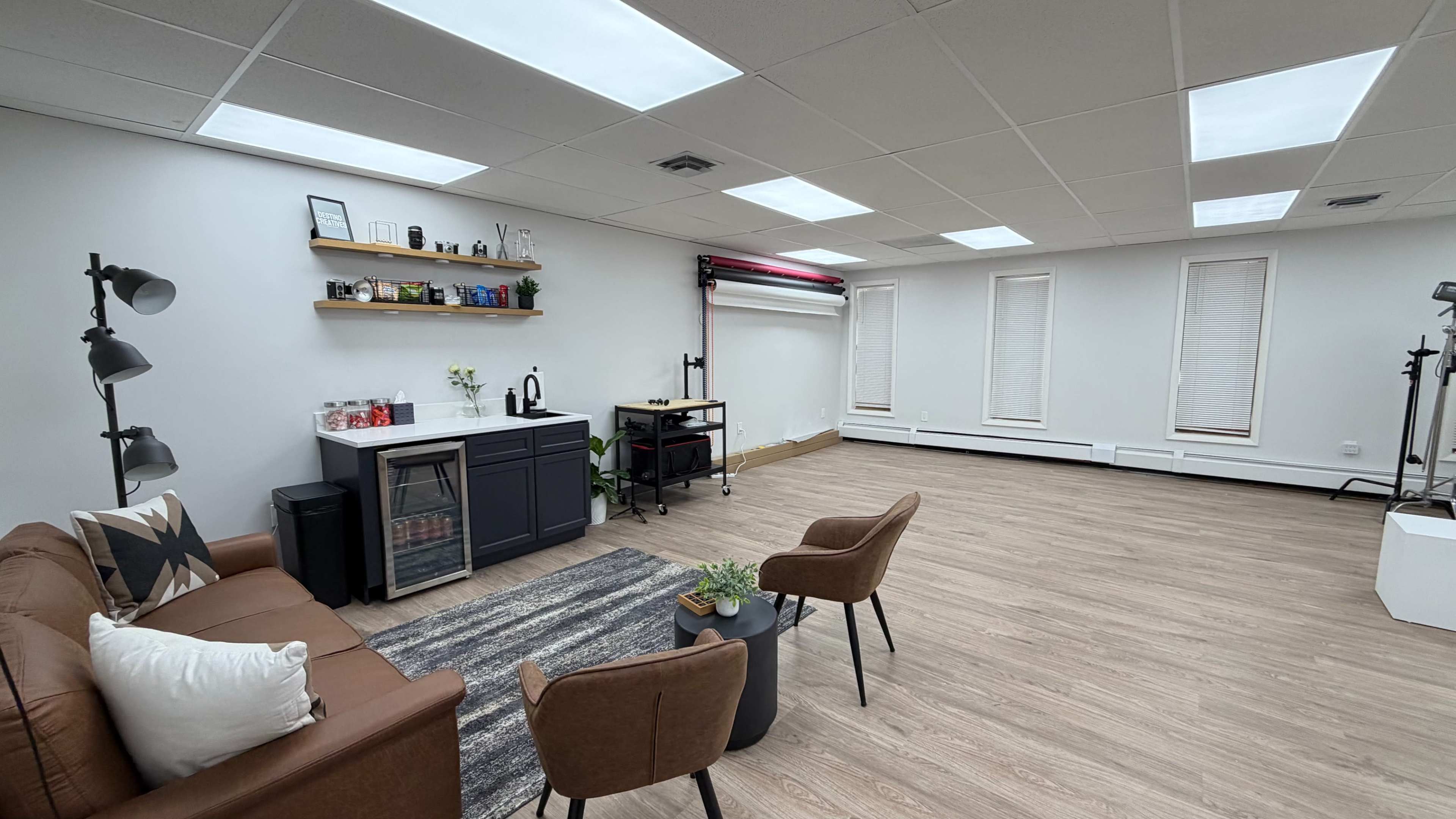 The image shows a spacious, well-lit room with a sofa, two chairs, a small table, and a kitchenette along one wall, featuring a shelf with various items.