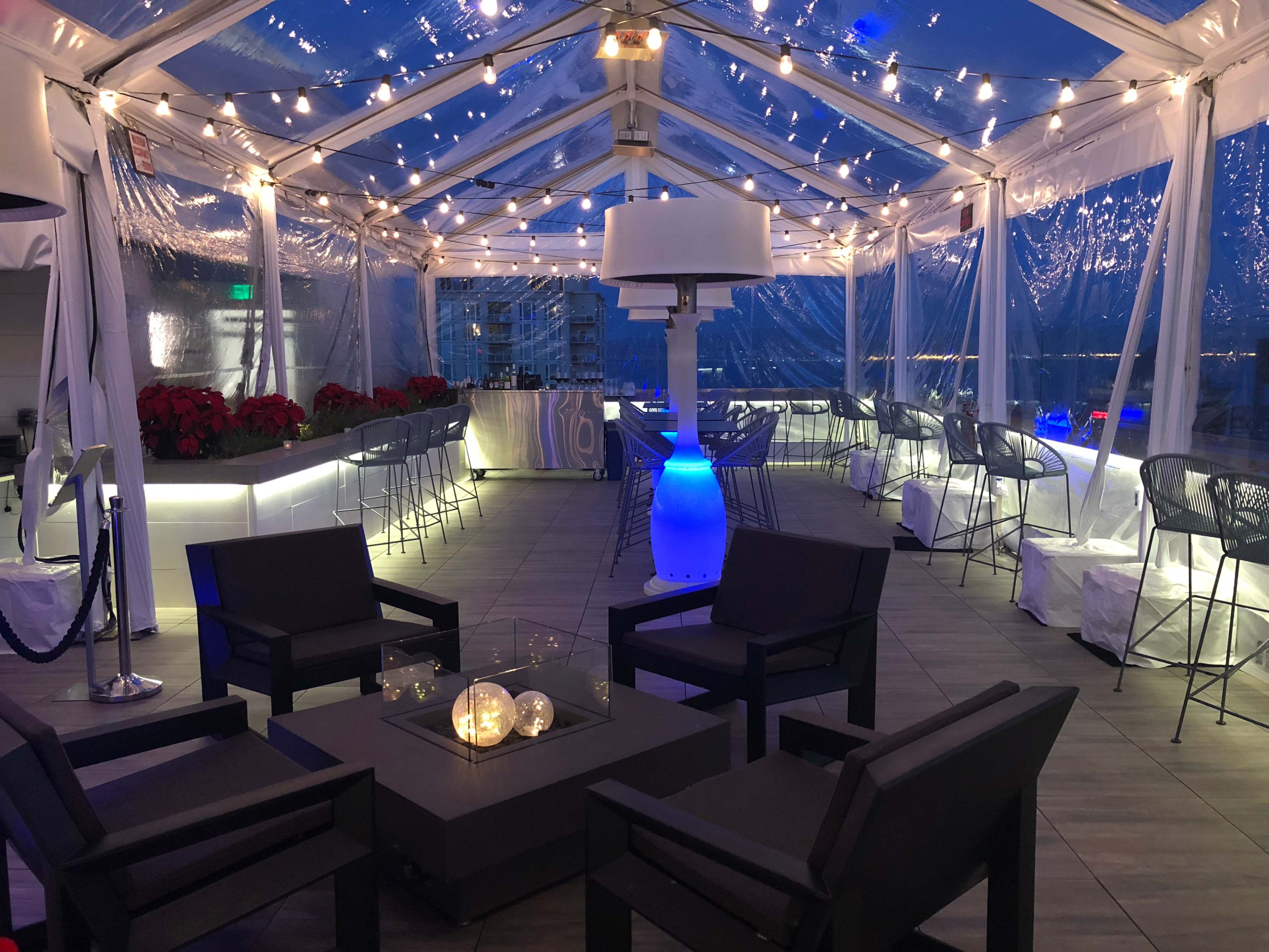 A covered outdoor lounge area features modern seating, string lights, and a glowing blue lamp amidst a festive setting.