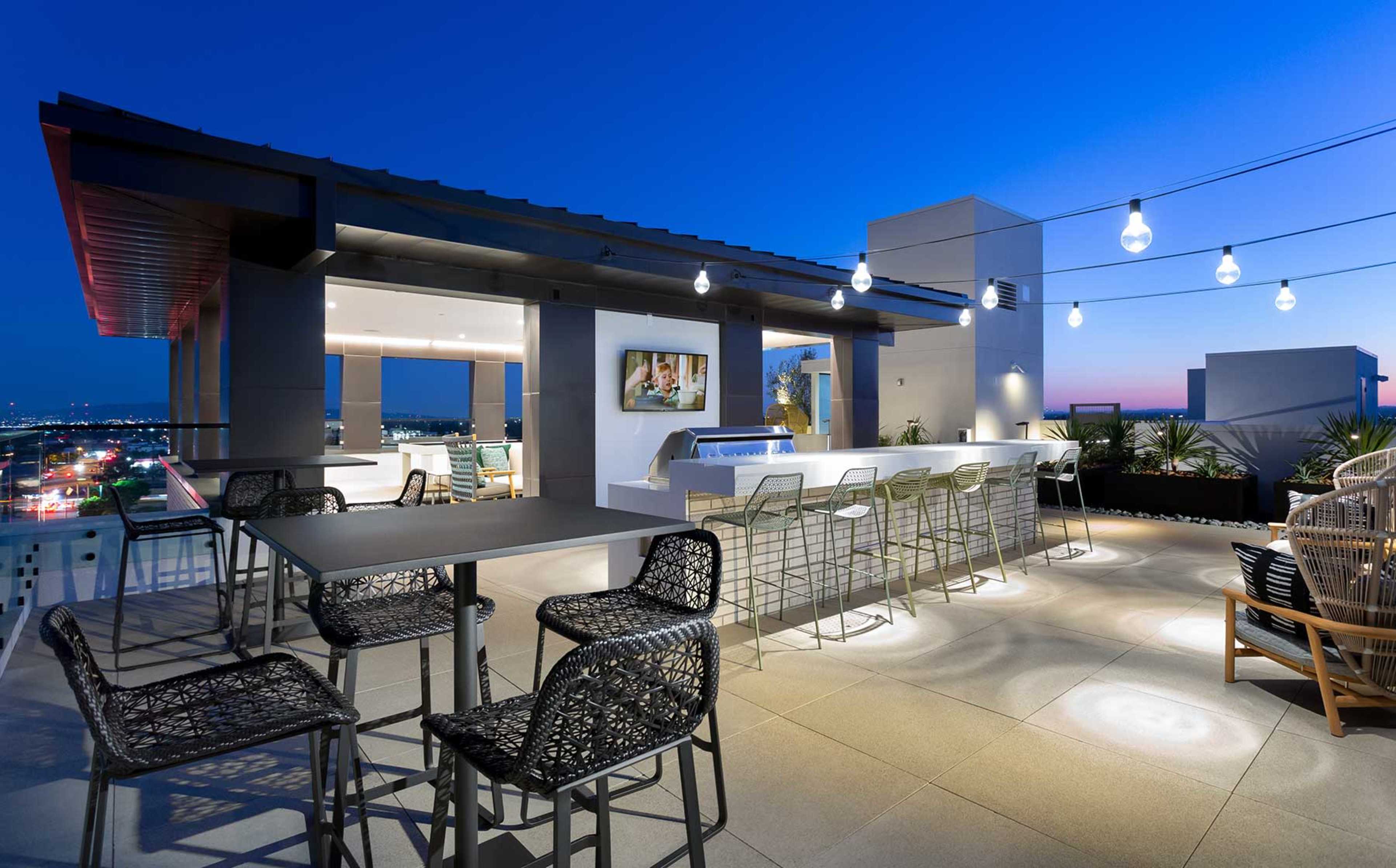 The rooftop patio features a bar area with high stools, string lights overhead, and a view of the city skyline at dusk.