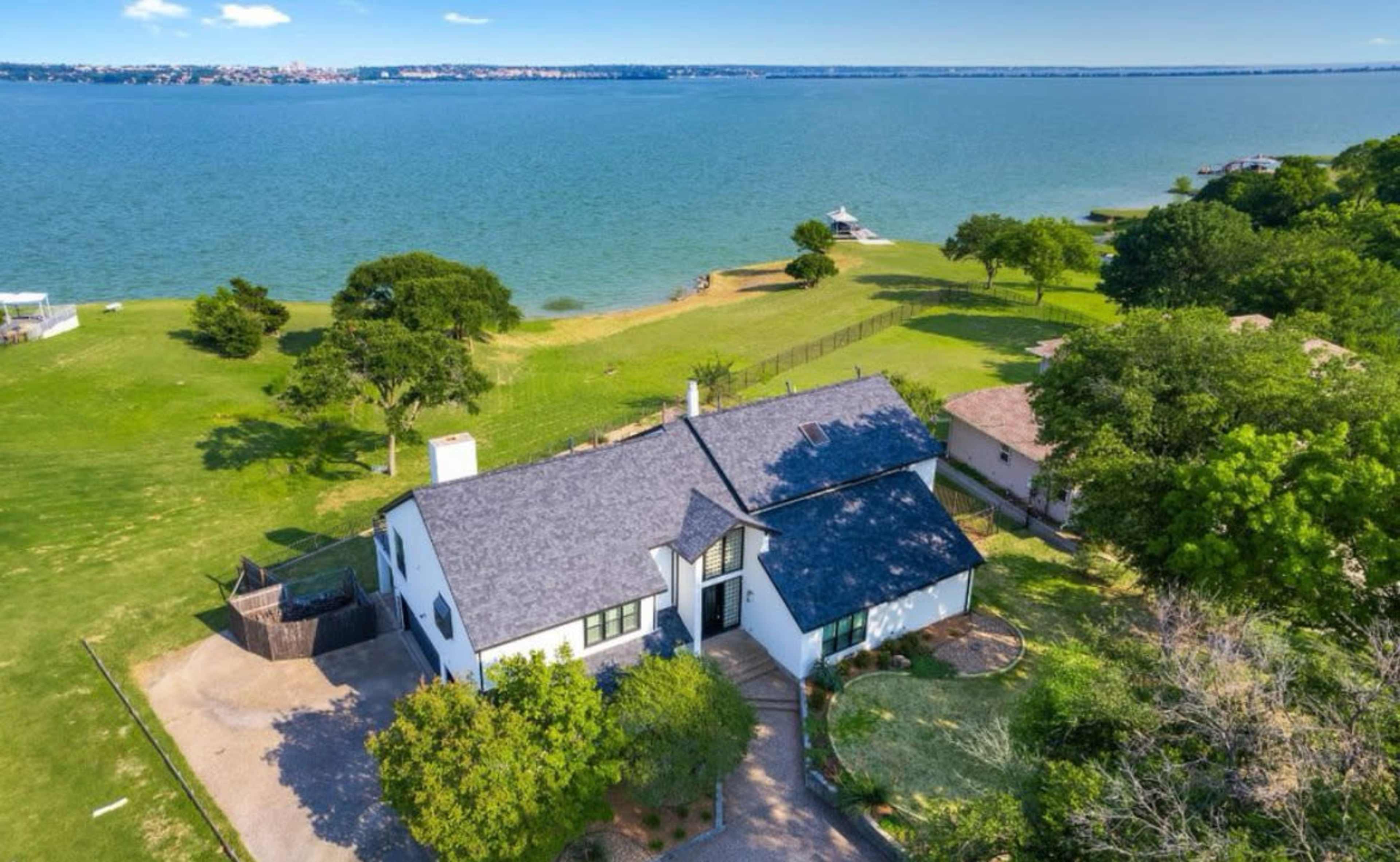 The image shows a waterfront house situated beside a lake, surrounded by green lawns and trees.