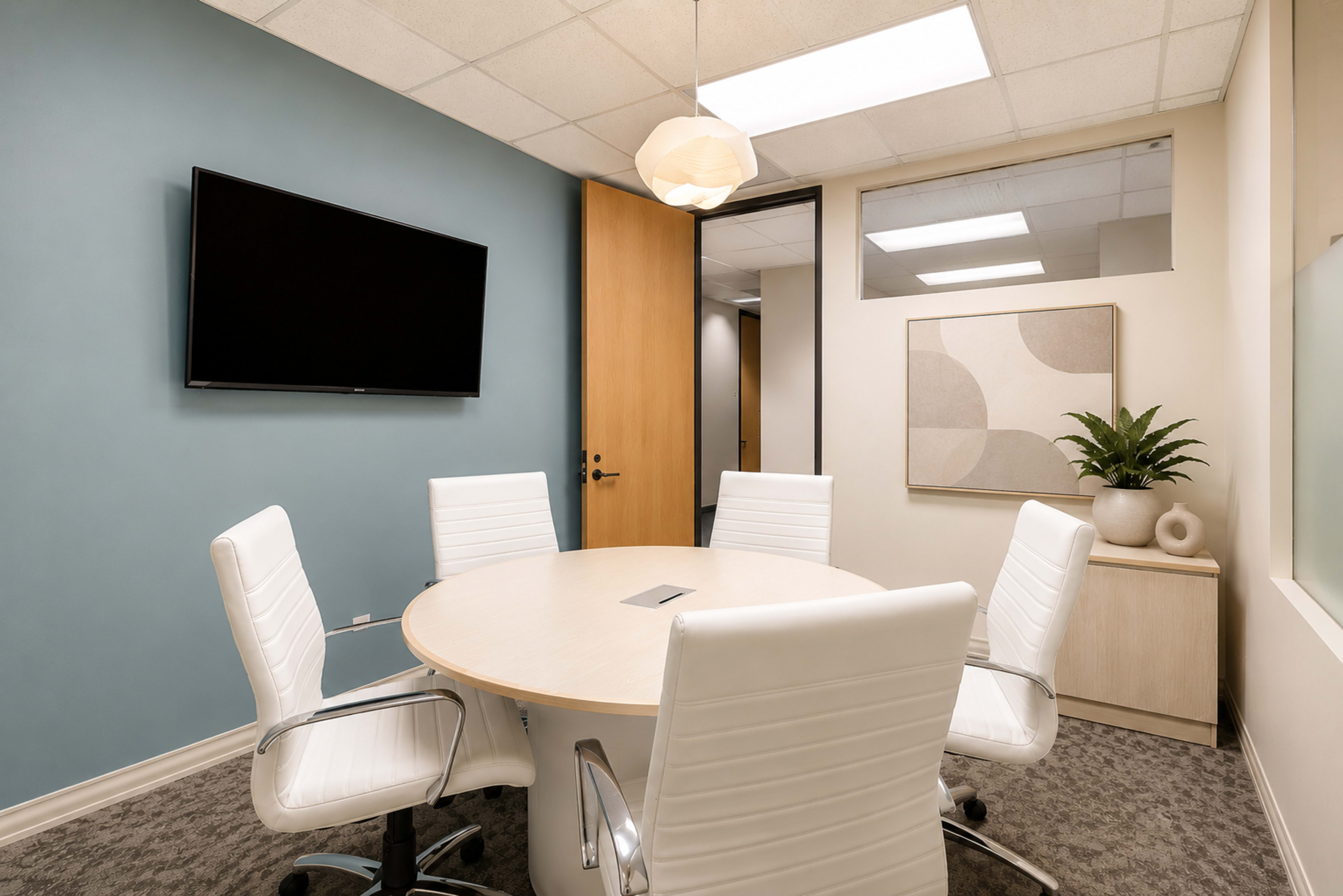 A modern conference room features a round table surrounded by white chairs, with a wall-mounted television and a potted plant.