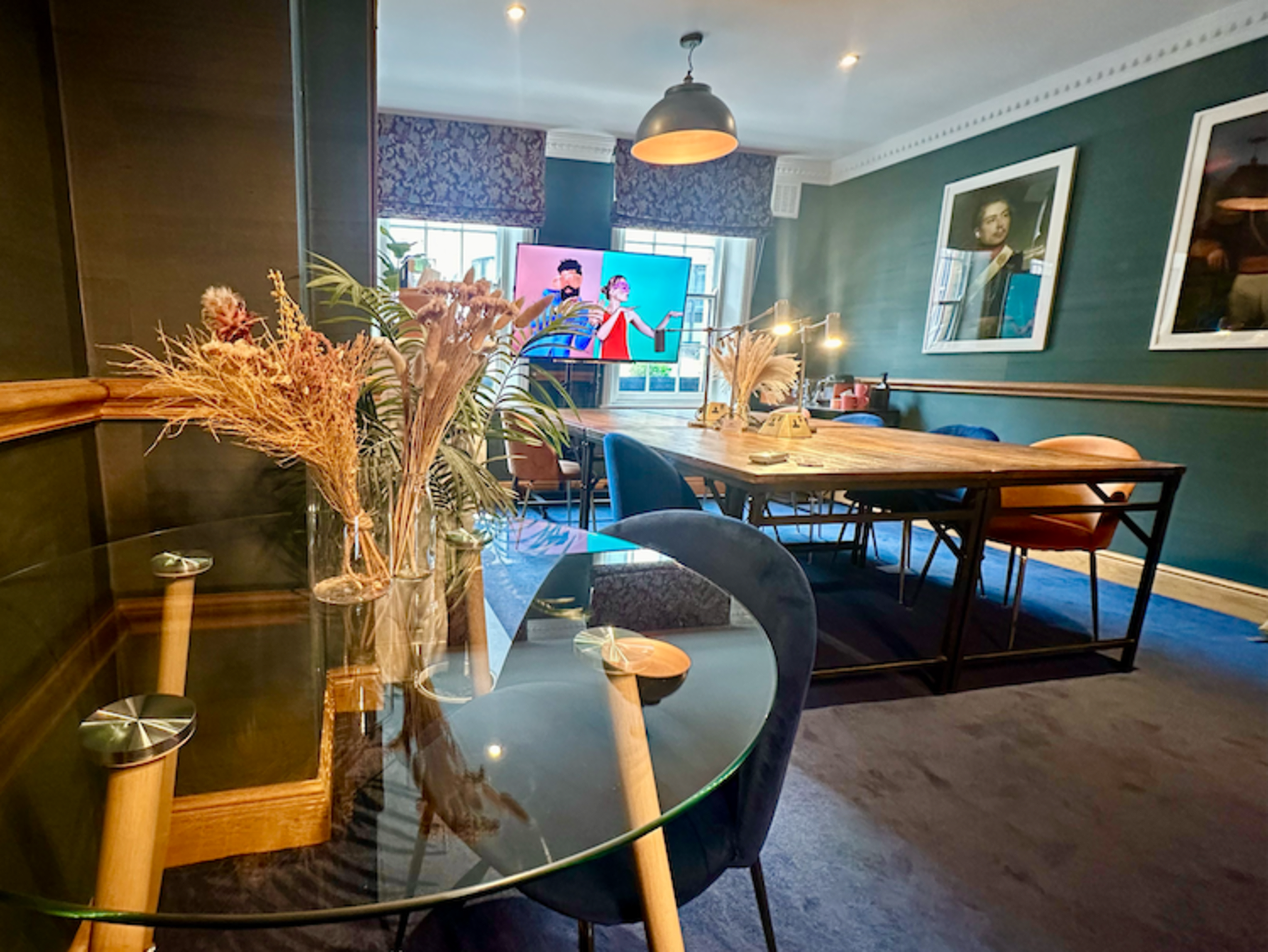 The image shows a stylish dining area featuring a large wooden table, modern chairs, and a television displaying colorful content, all set against dark green walls and decorative accents.