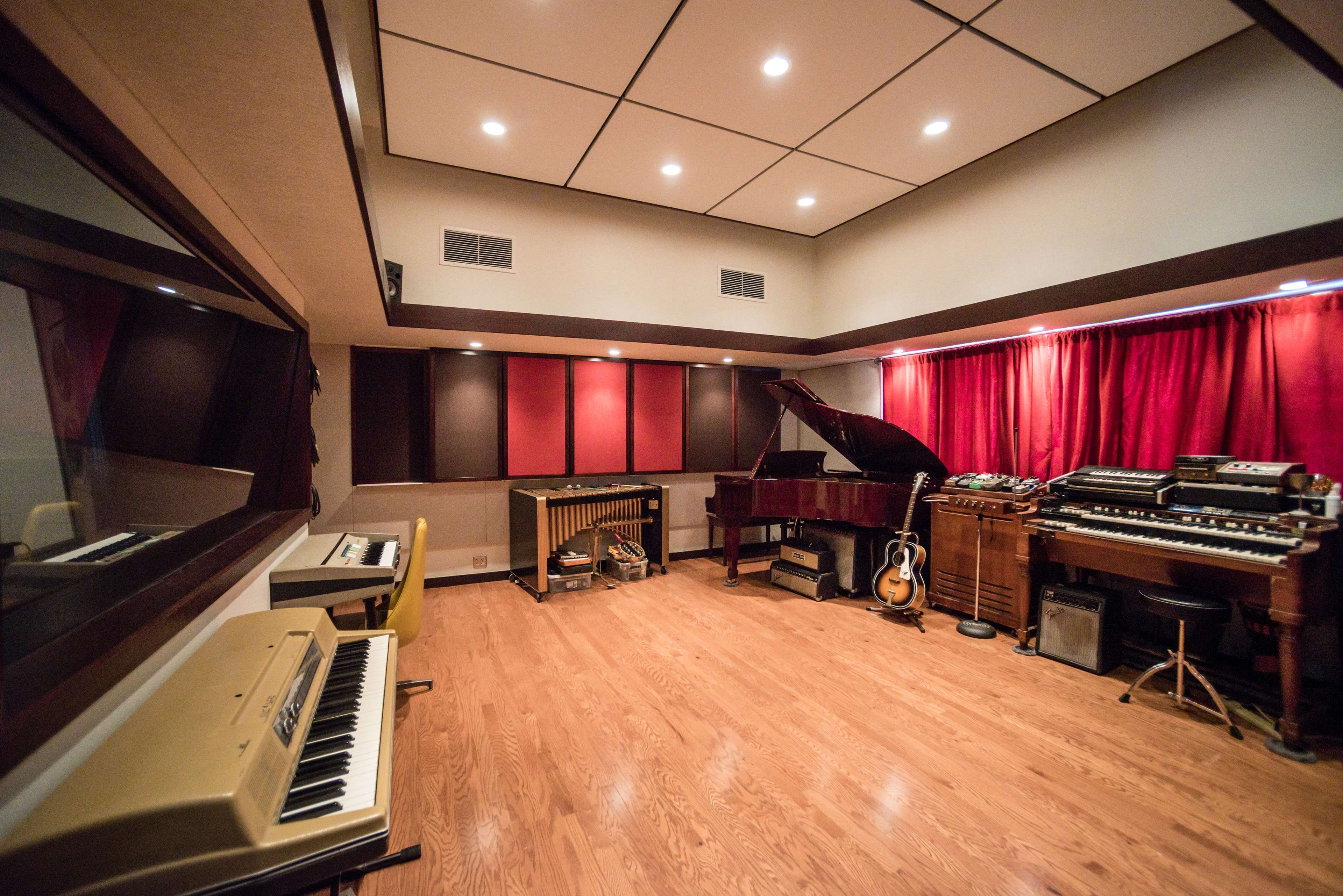 The image shows a music studio featuring a grand piano, various keyboards, a vibraphone, an electric guitar, and several vintage audio equipment arranged in a well-lit room with red curtains.