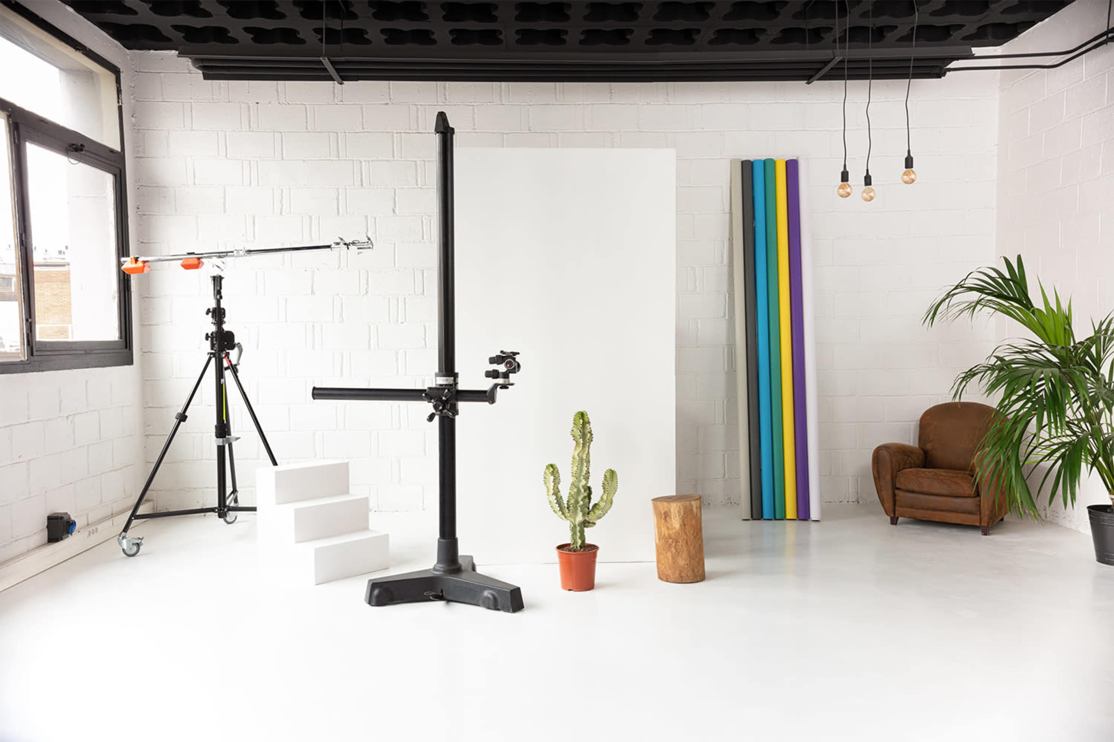 A bright, spacious studio with a white backdrop, a cactus in a pot, a wooden stool, a camera setup, and colorful vertical panels.