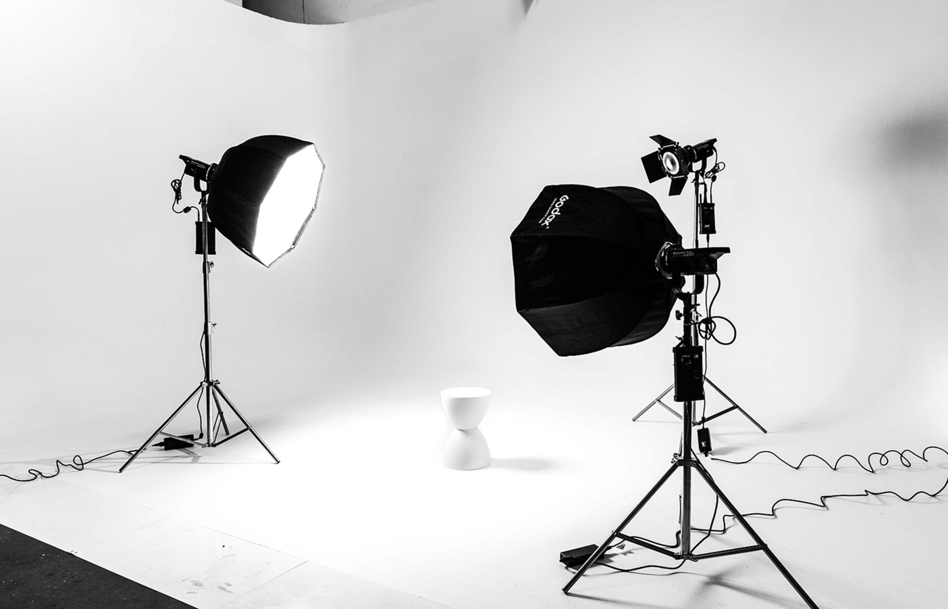 A photography studio setup with two light sources and a single white stool positioned in front of a plain backdrop.