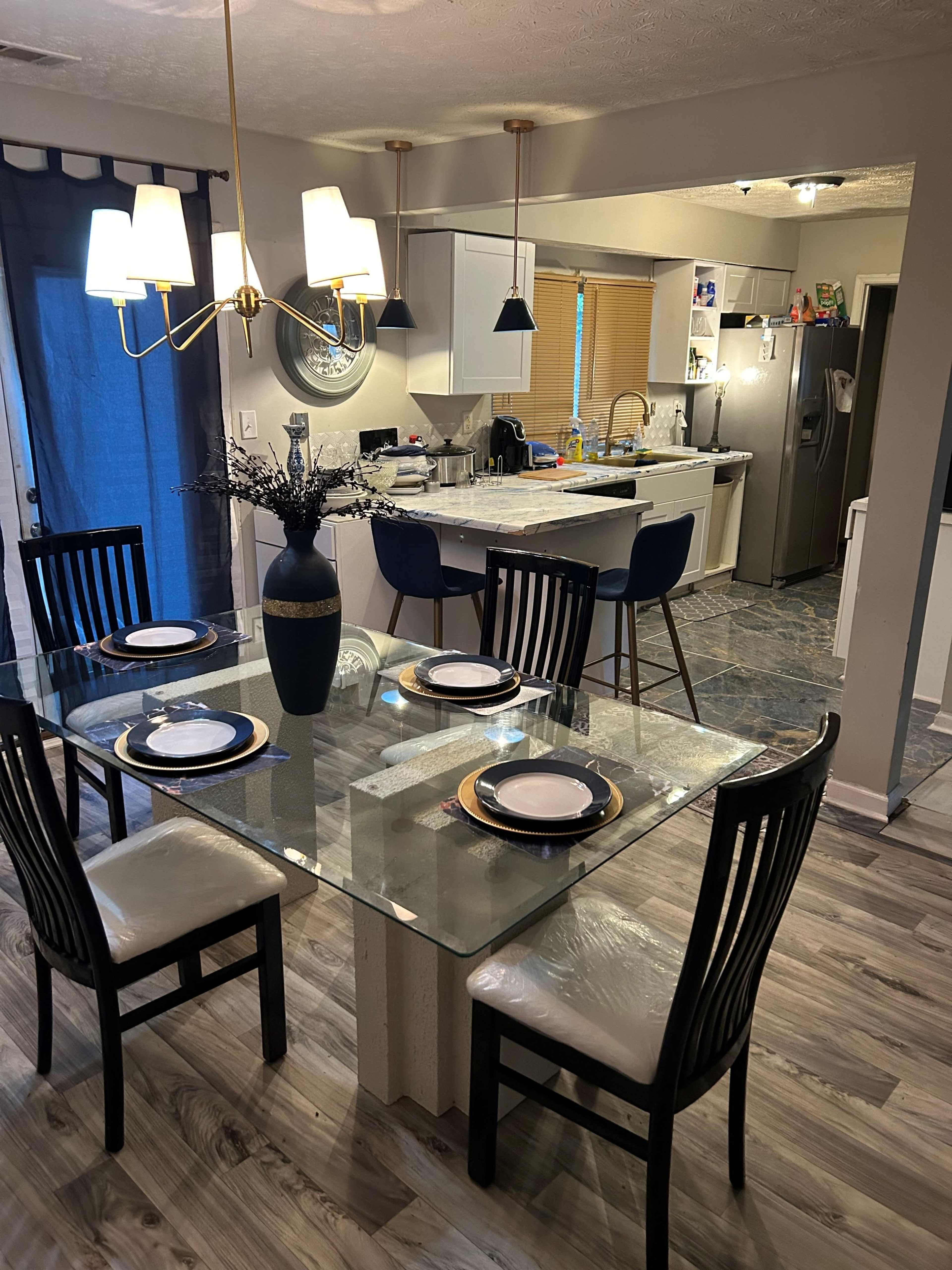A modern dining area features a glass table set for four, with a kitchen and living space visible in the background.