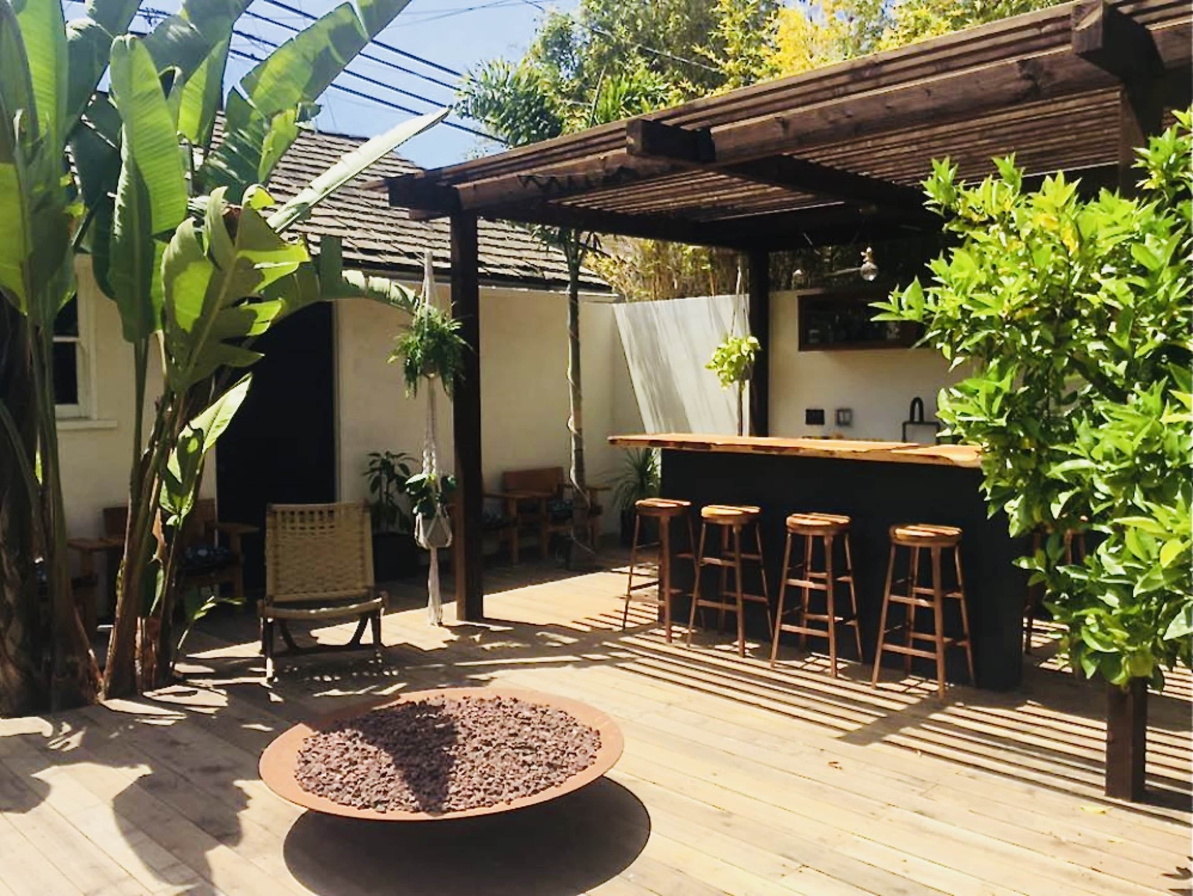 A wooden deck with outdoor seating, featuring a bar area and a circular fire pit surrounded by plants.
