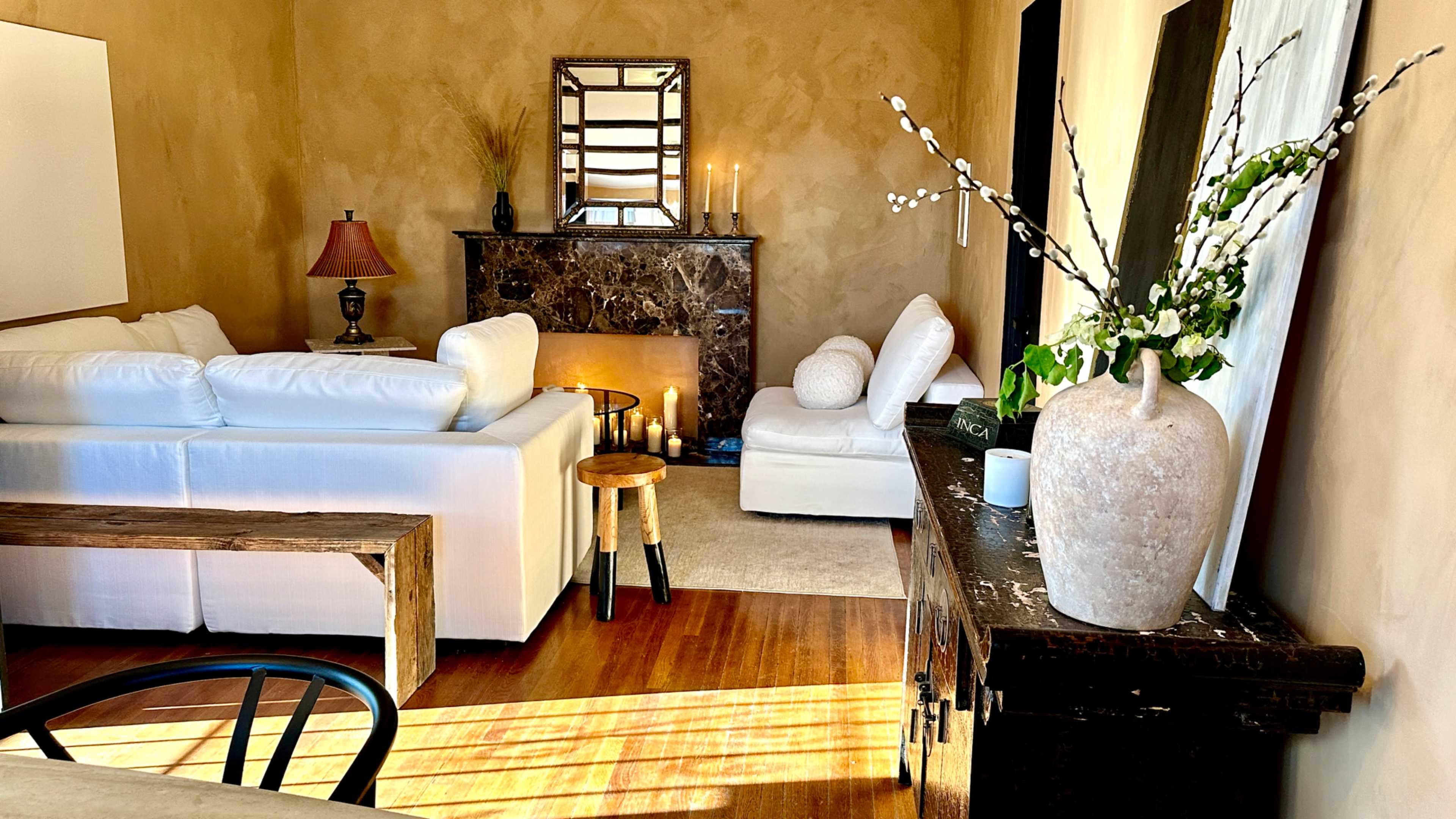 The image shows a neatly arranged living room with a white sofa, a marble fireplace adorned with candles, and a decoratively styled table featuring a vase and branches.