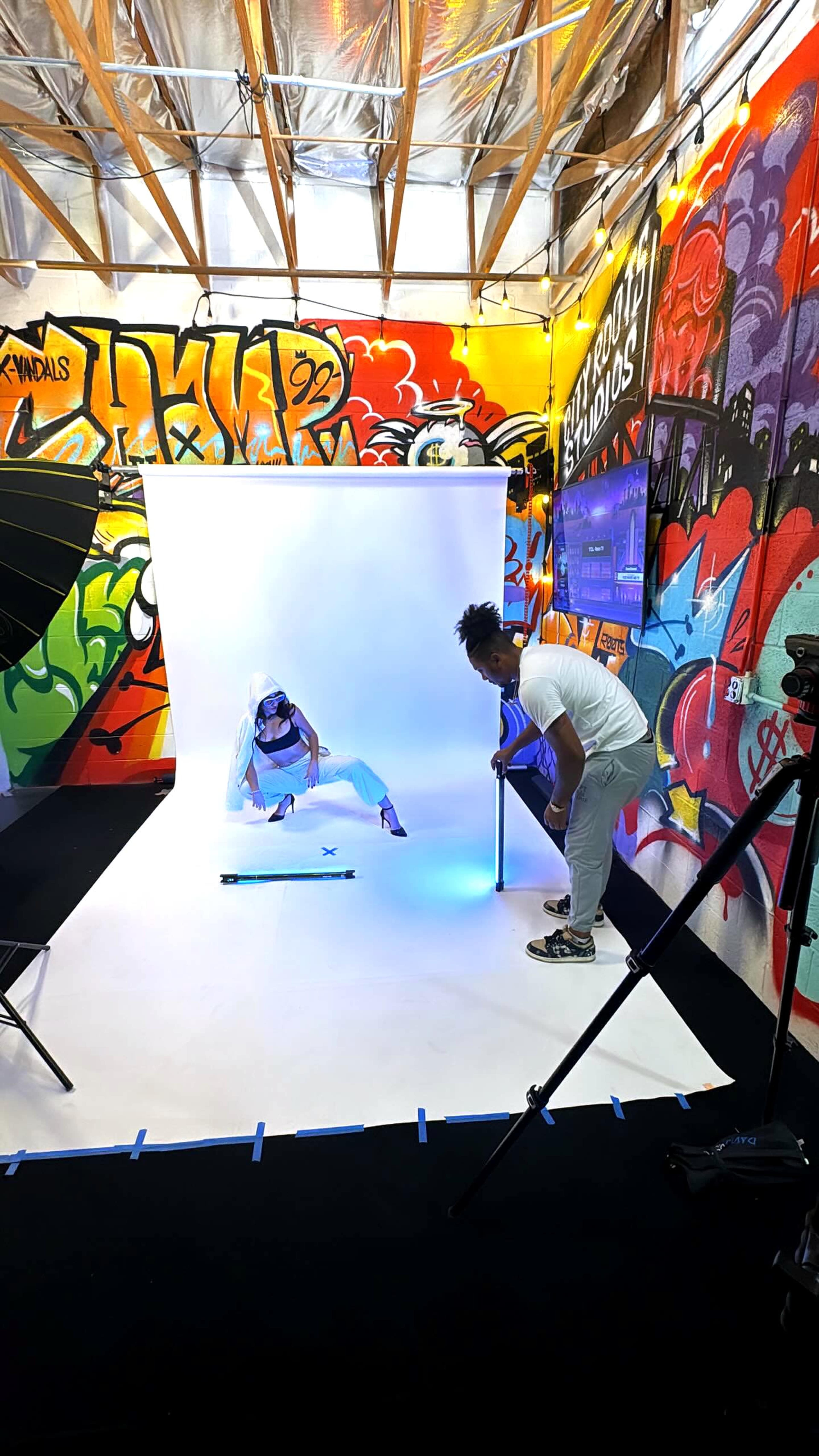 A person poses in a studio with a white backdrop while another adjusts equipment in front of colorful graffiti-covered walls.