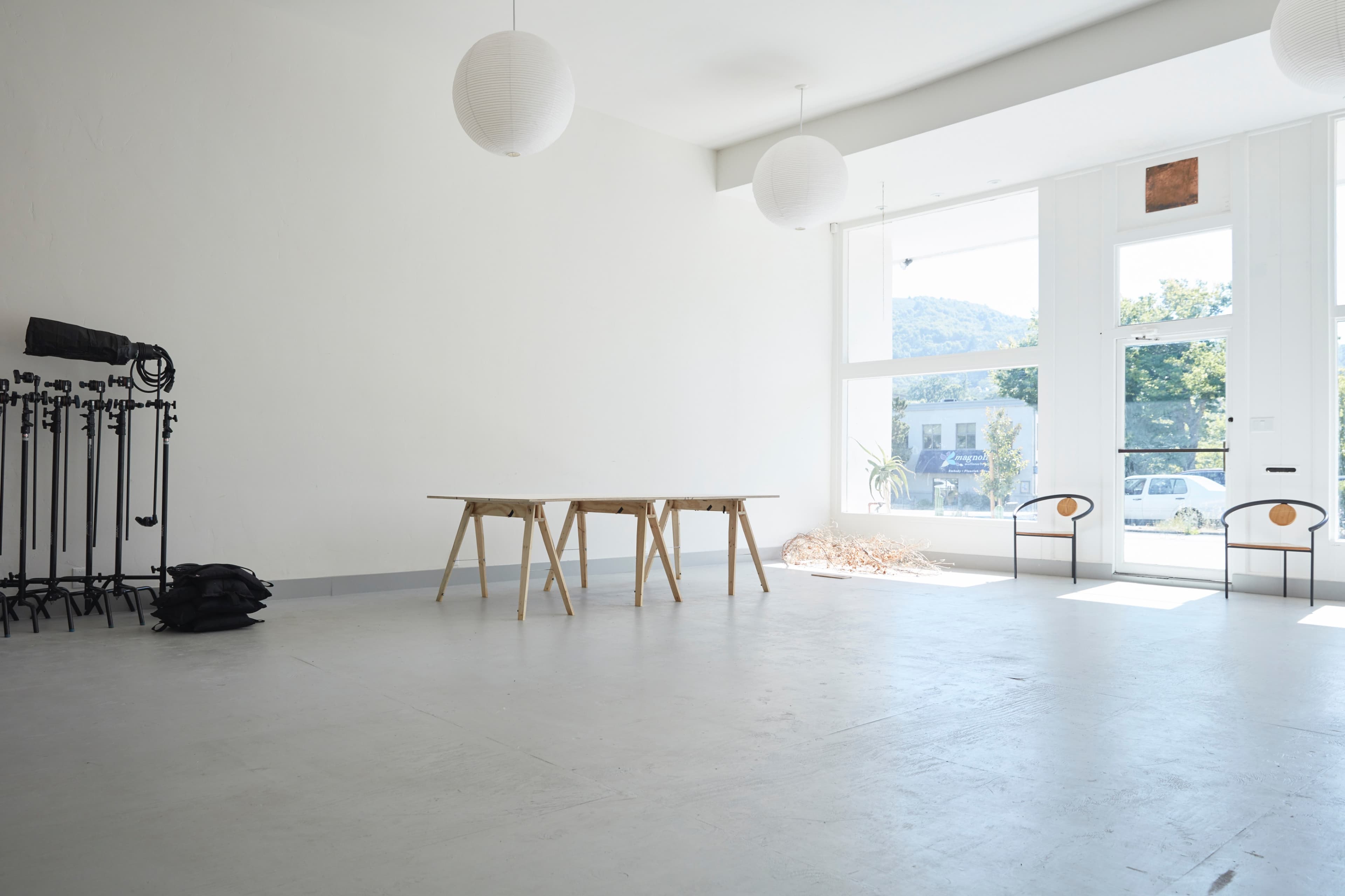The image shows a spacious, empty room with large windows, two chairs, a wooden table, and lighting equipment on the side.