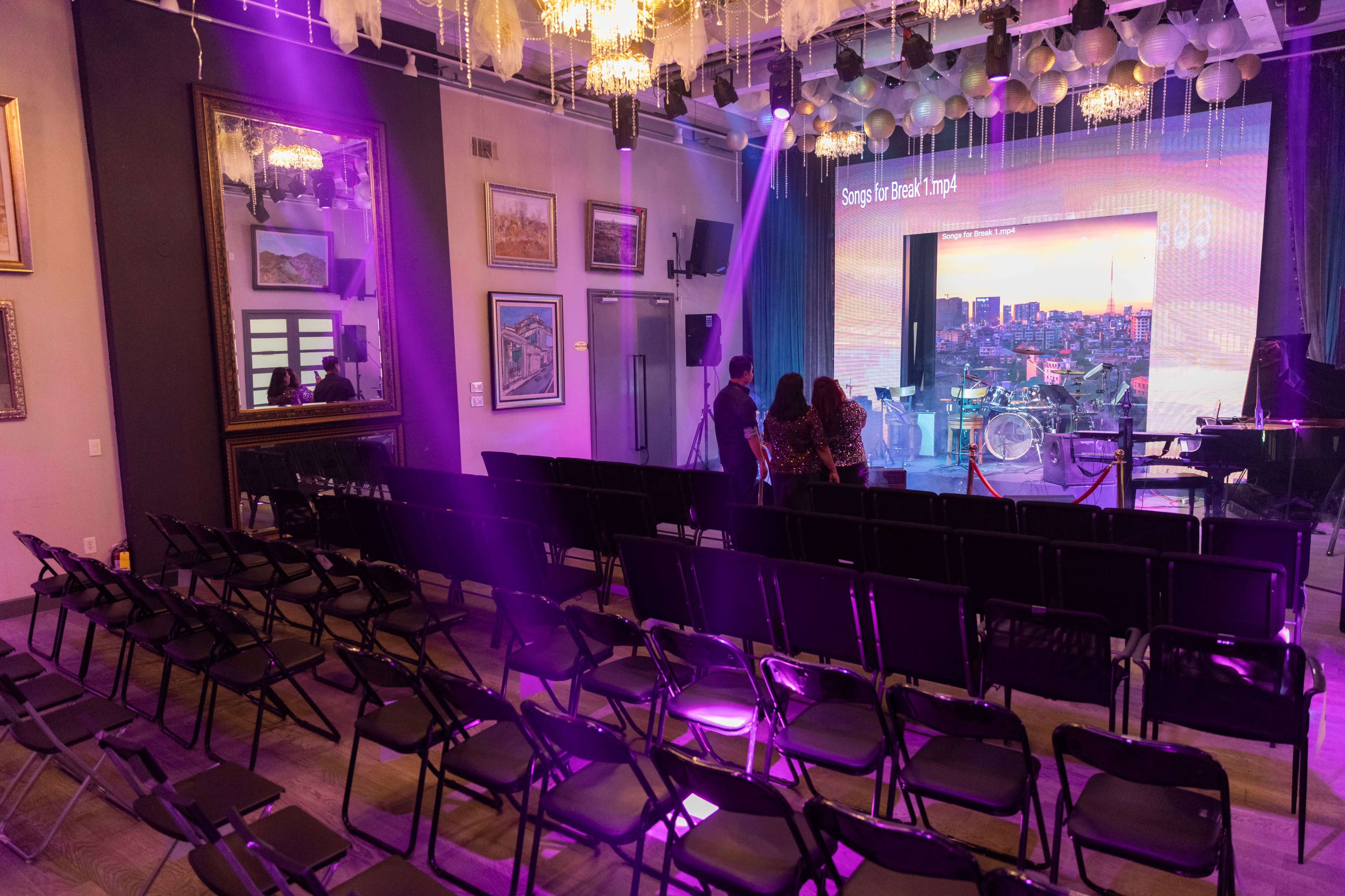 The image shows a performance venue with rows of empty chairs, a stage set up with a backdrop displaying a cityscape, and purple lighting illuminating the space.