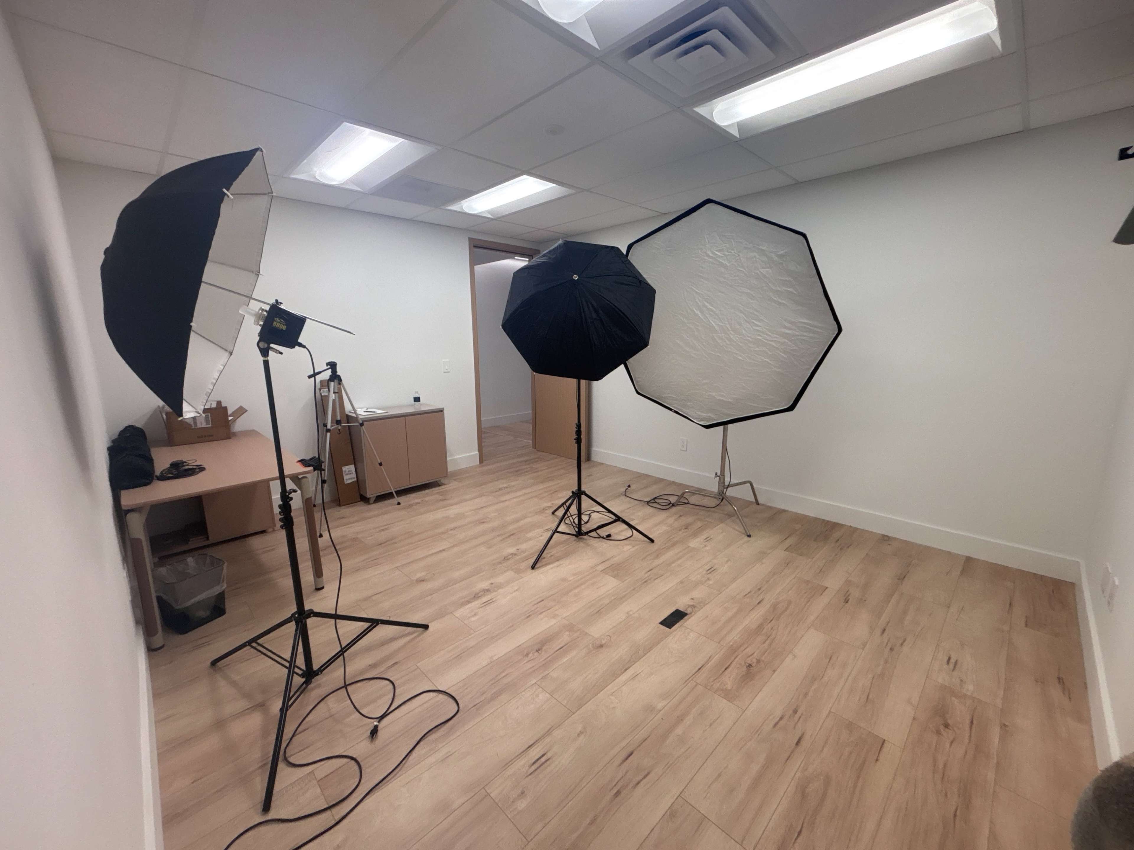 The image shows a small photography studio equipped with two light umbrellas, a tripod, and a worktable against a plain wall.