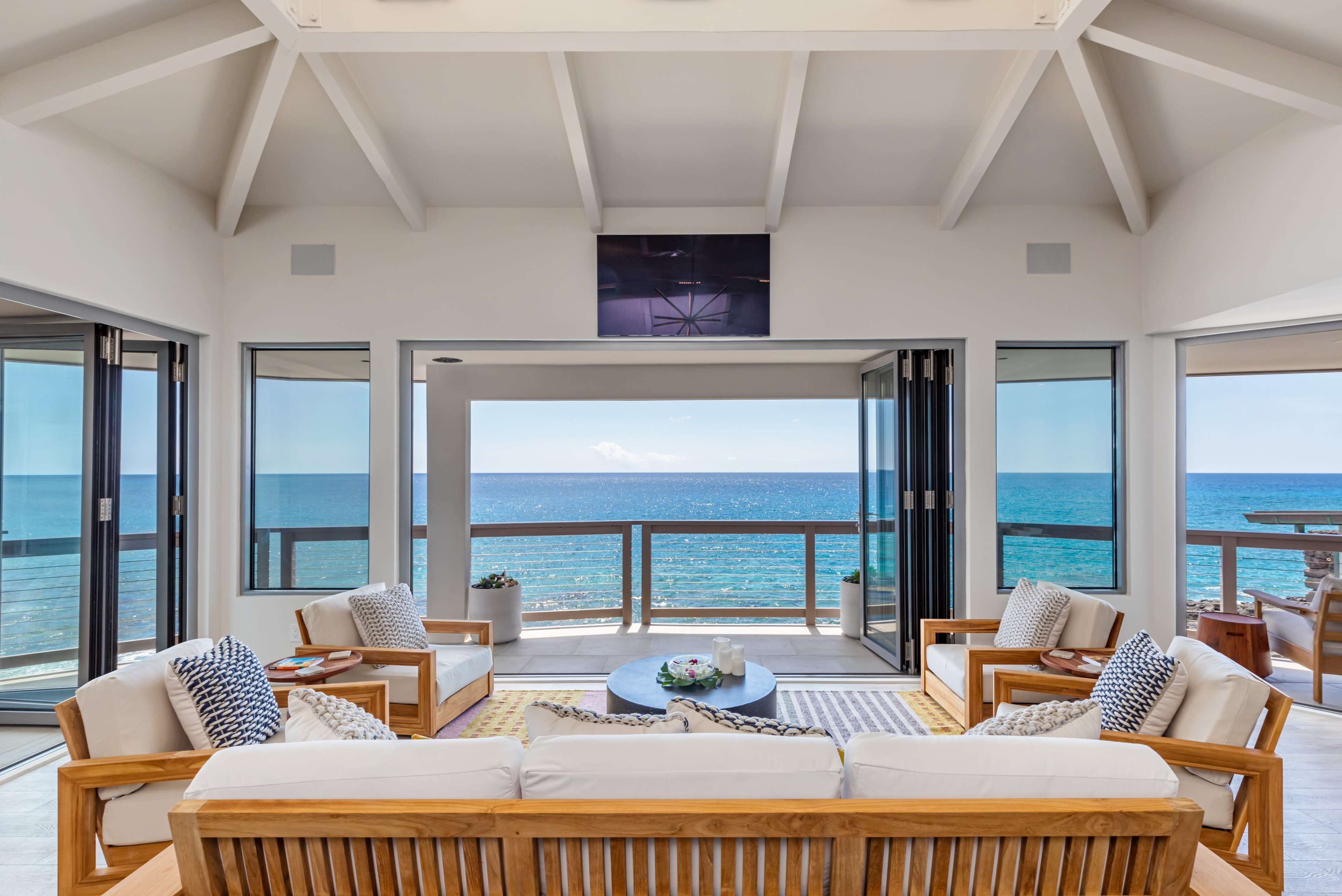 A coastal living room features large glass doors opening to a panoramic ocean view, with wooden furniture arranged around a central coffee table.