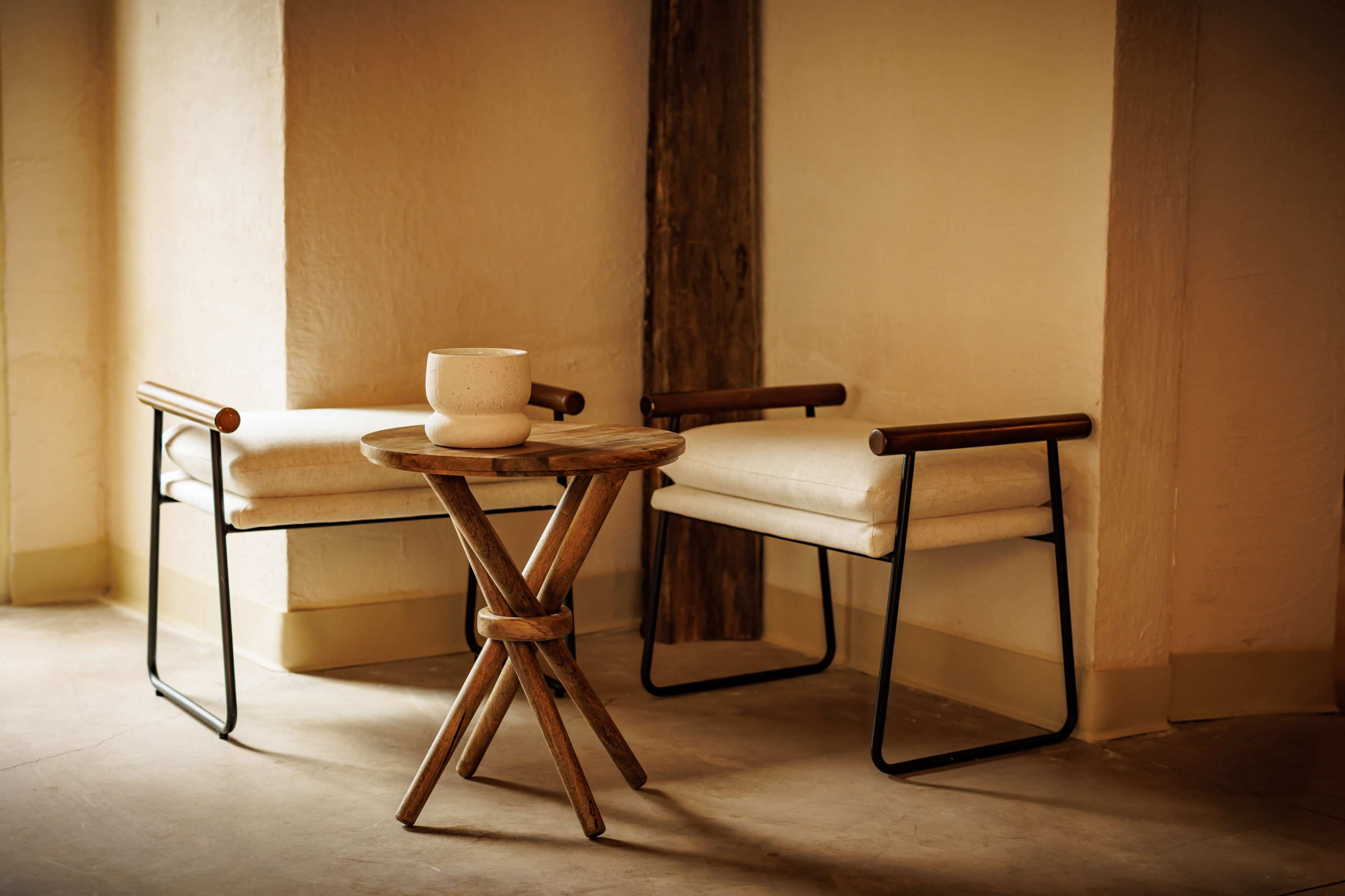 Two modern chairs with fabric cushions are positioned beside a wooden tripod table holding a ceramic pot in a softly lit corner.