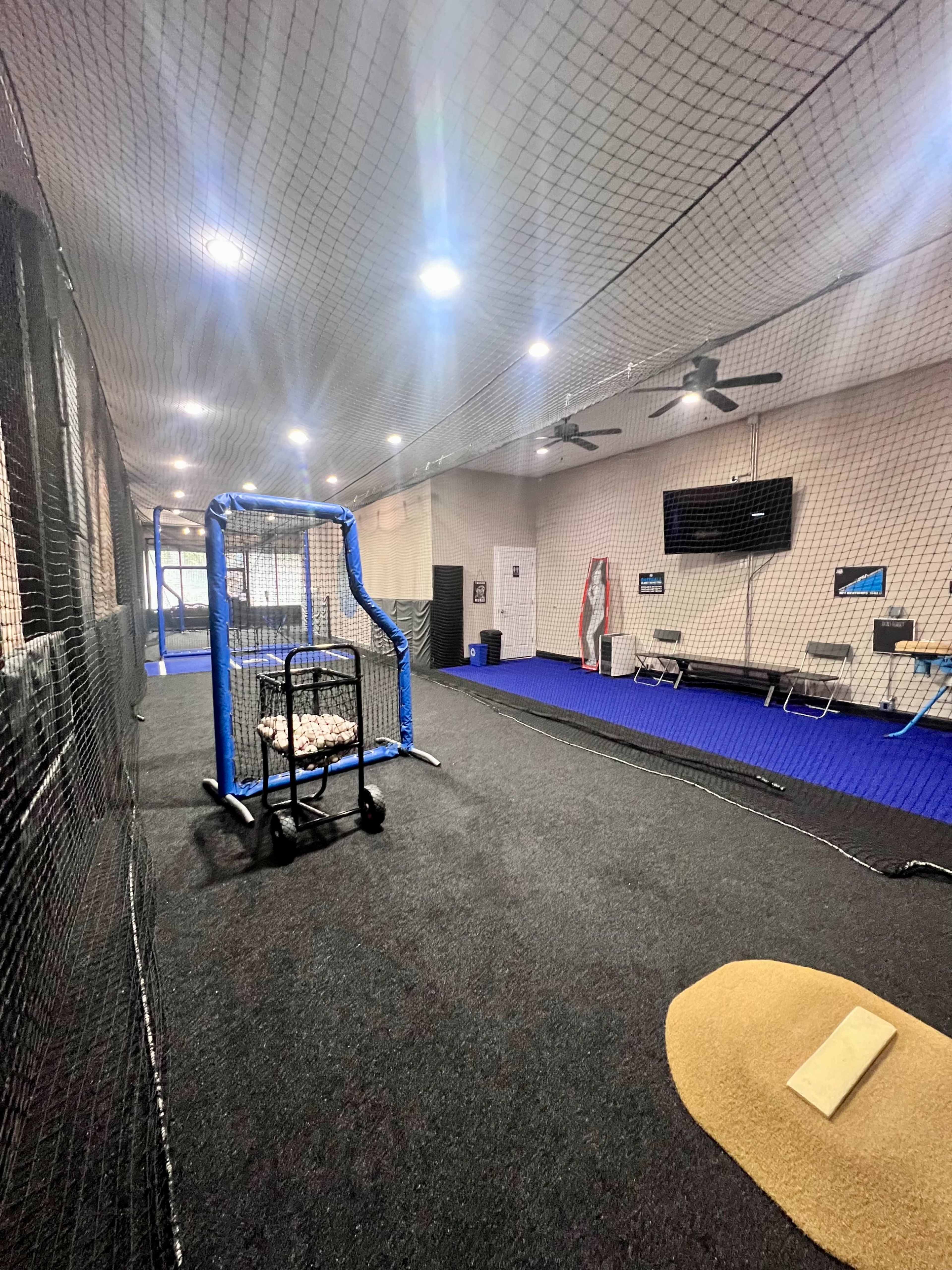 An indoor baseball training facility featuring a batting cage, a pitching area, and various training equipment on a synthetic turf floor.