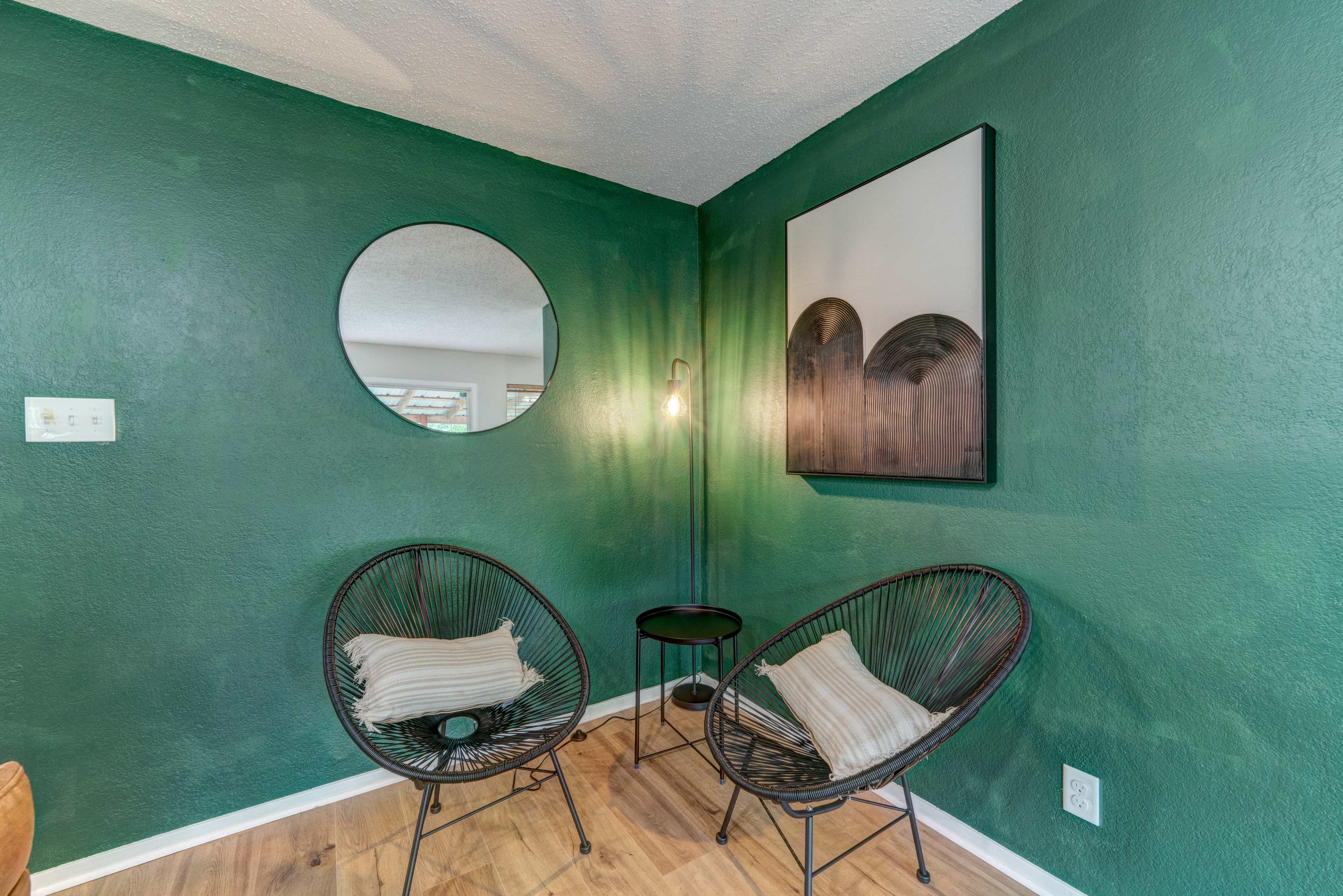 A small corner with two wire chairs, a round mirror on the wall, a lamp, and a circular table, all set against a green-painted wall.