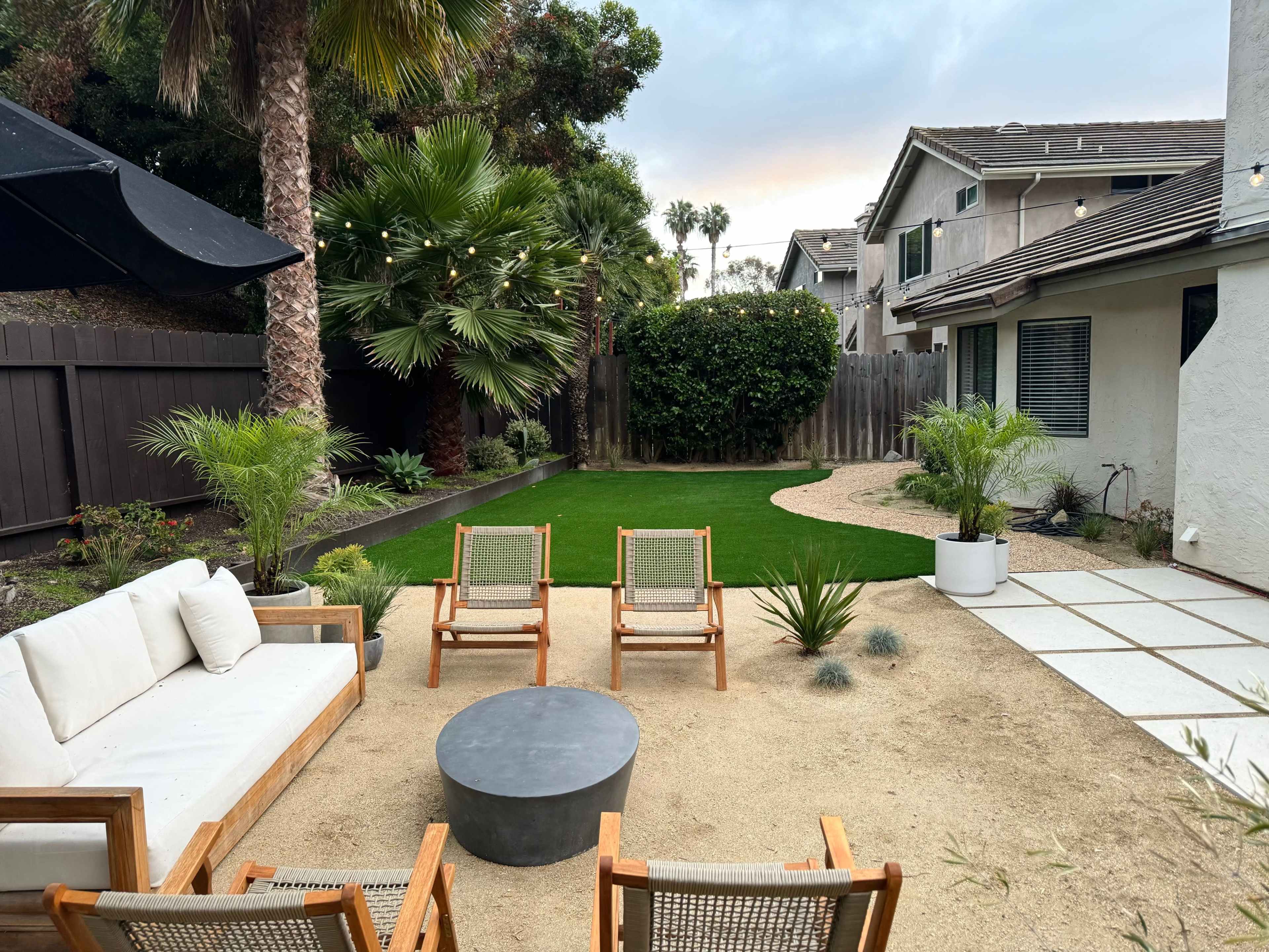 A landscaped backyard featuring a seating area with three wooden chairs, a round gray table, and green grass bordered by plants and a pathway.