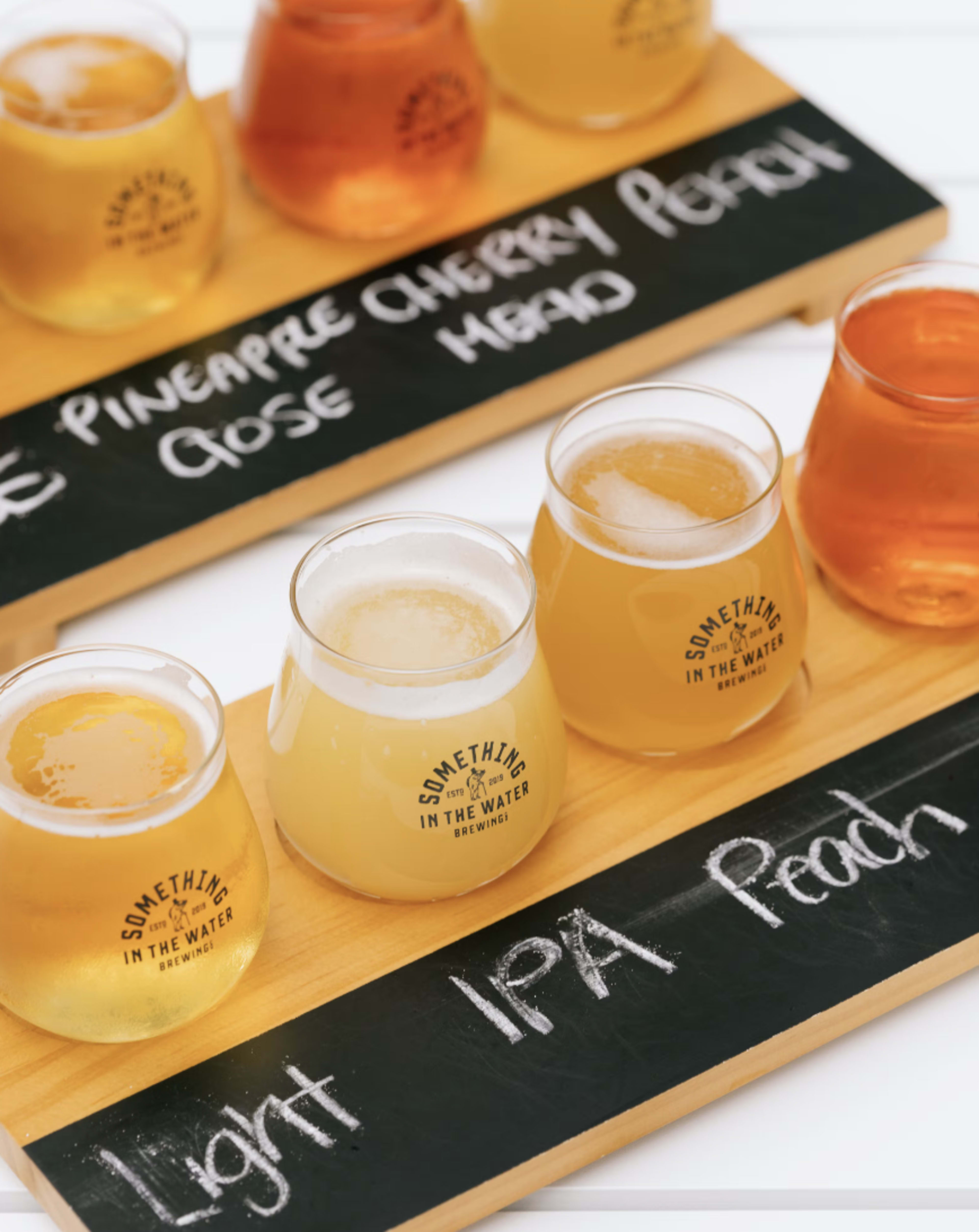 A wooden tasting board displays several glasses of craft beer, each labeled with the names of the different varieties including "Light," "IPA," and "Peach."