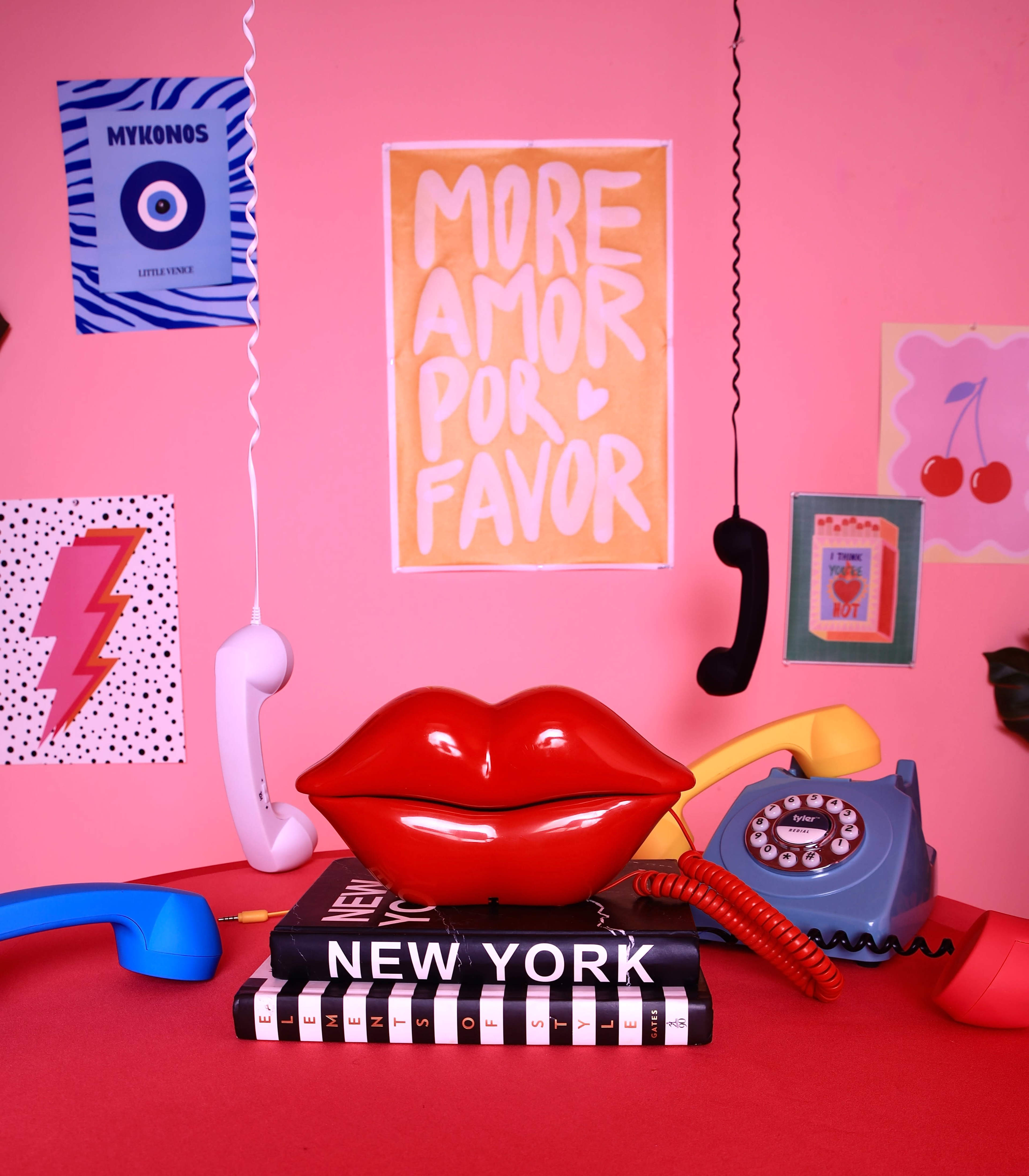 The image features a colorful tabletop scene with a red lip-shaped decor item, several vintage-style phones, and various printed artworks against a pink background.