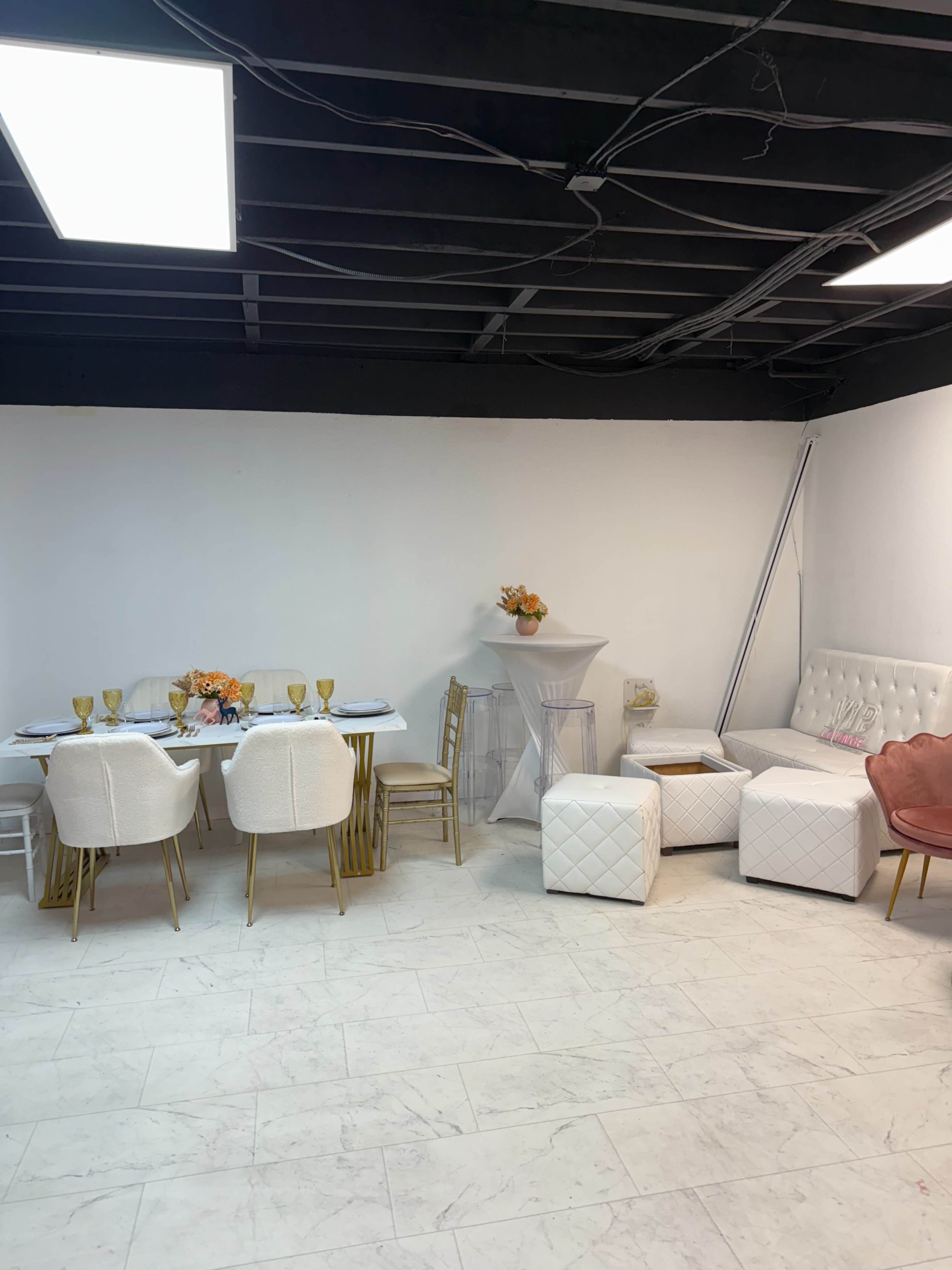 The image shows a minimalist event space featuring a white table set with gold utensils, white chairs, a small round table with a floral arrangement, and various seating options in the background.